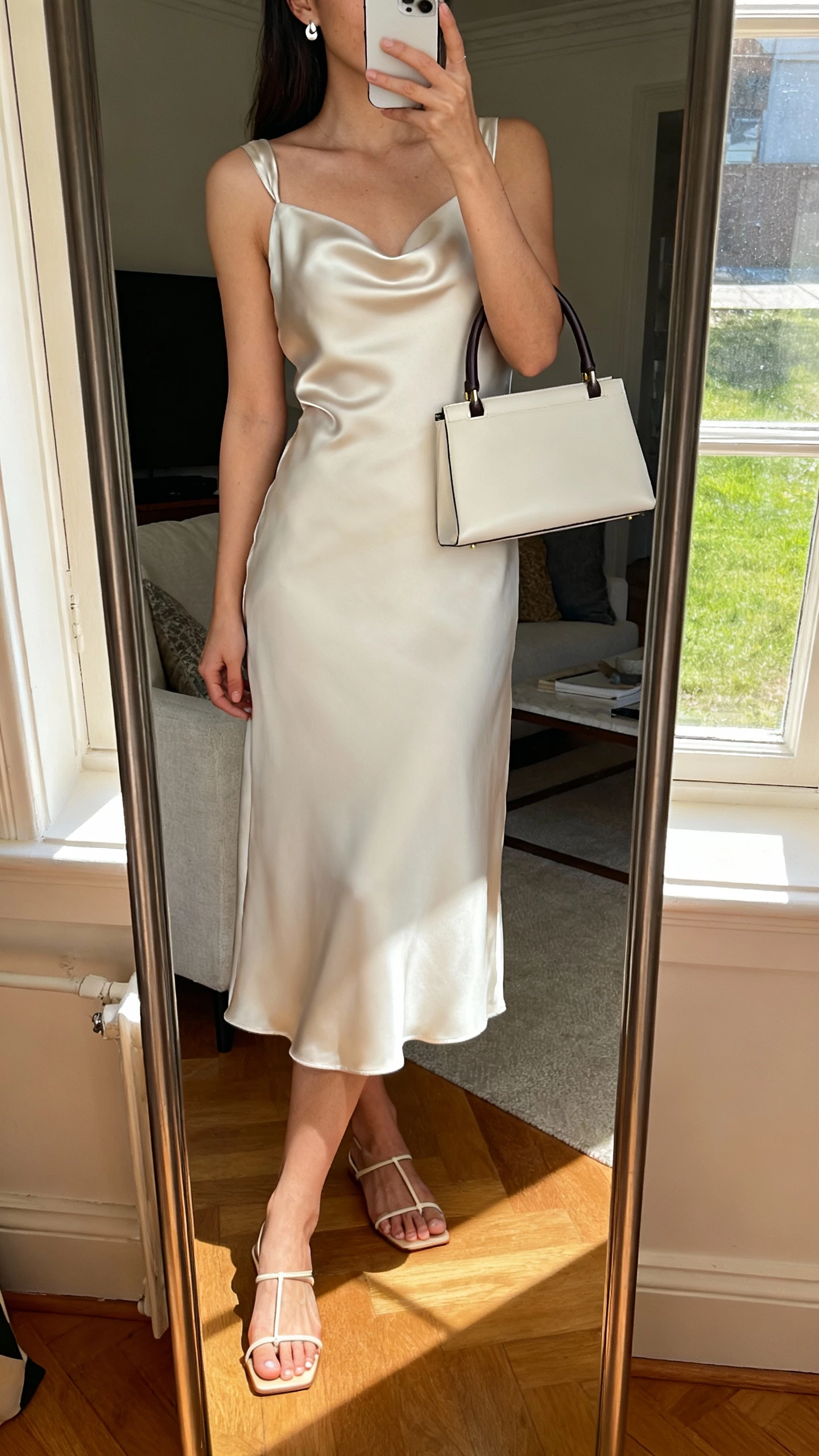 iPhone mirror selfie of a woman wearing a satin-back crepe midi dress with clean minimalist lines, barely-there sandals, simple stud earrings, and a structured top-handle bag, face not visible, living room mirror, daylight from window, natural iPhone quality.