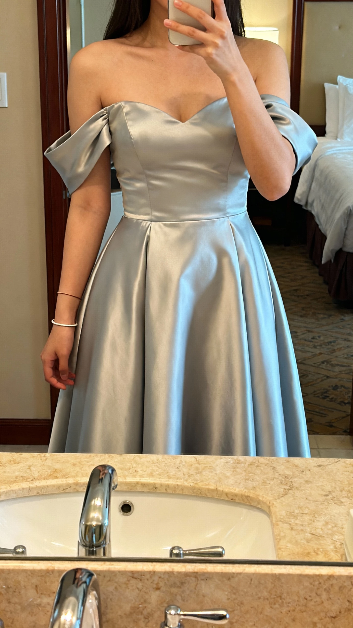 iPhone mirror selfie of a woman wearing a satin fit-and-flare gown with detachable off-the-shoulder sleeves, thin bracelet, face not visible, hotel bathroom mirror, warm indoor lighting, natural iPhone photo quality.