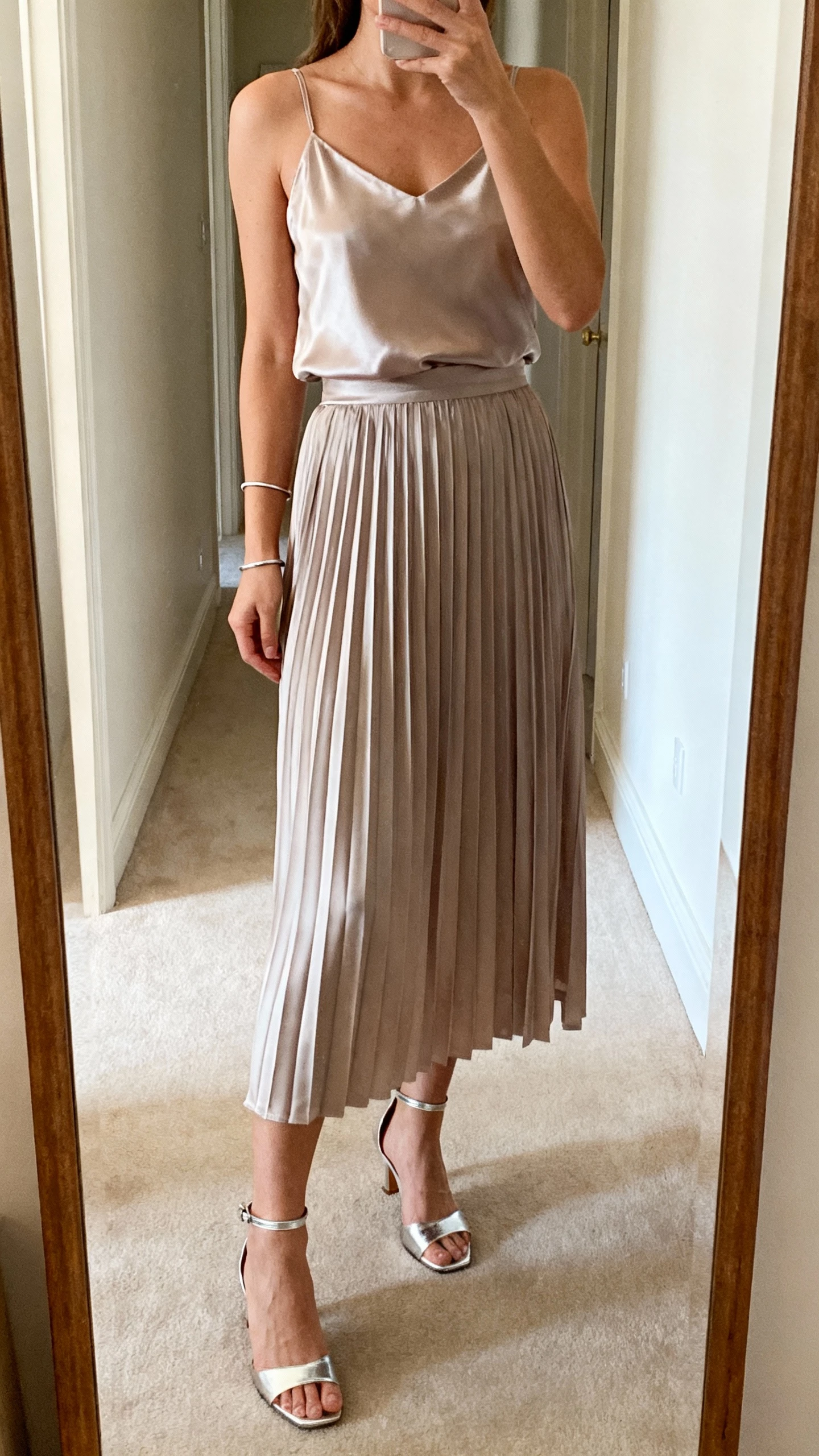 iPhone mirror selfie of a woman wearing a satin slip-top and a pleated midi skirt set, metallic ankle-strap heels, slim bracelet, face not visible, full-length hallway mirror, indoor morning light, natural iPhone photo.