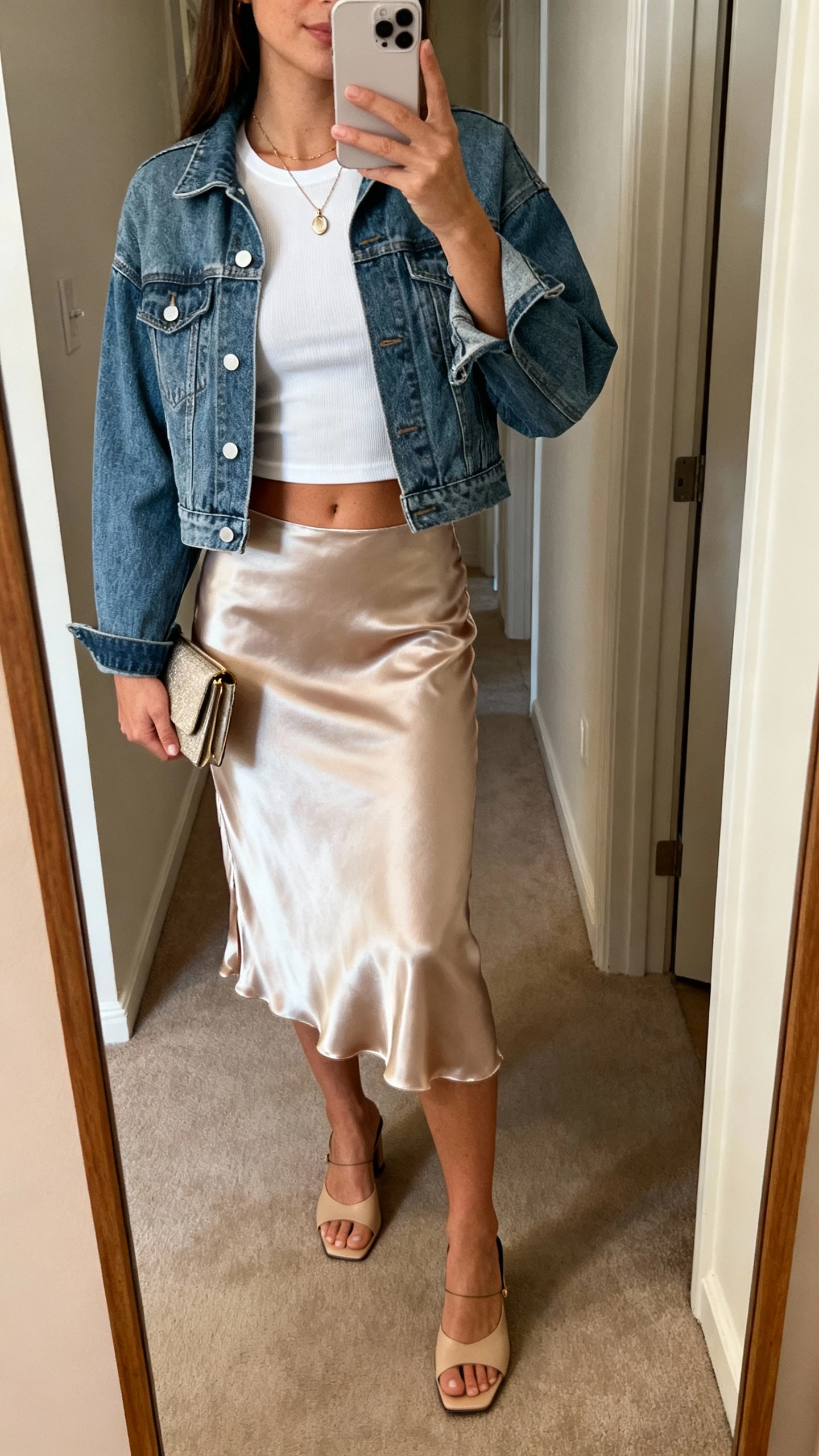 iPhone mirror selfie of a woman wearing a satin slip skirt, fitted tee tucked in, cropped denim jacket, delicate pendant necklace, low-heel mules, small clutch, face not visible, hallway mirror with warm indoor lighting, natural iPhone photo quality.