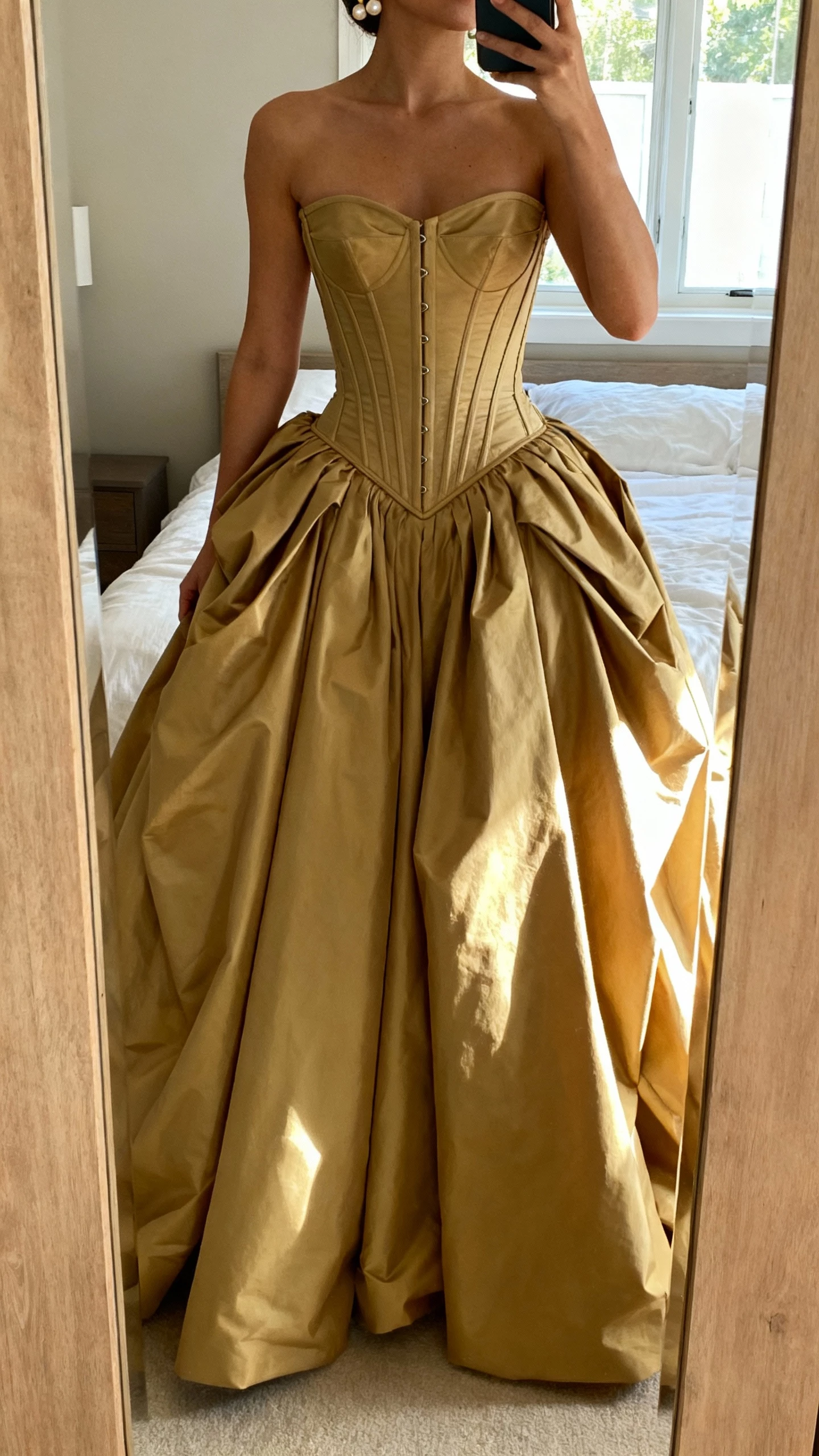 iPhone mirror selfie of a woman wearing a sculpted corset ballgown with structured bodice, dramatic full skirt, and supportive boning, minimal pearl earrings, face not visible, cropped at neck, bedroom full-length mirror, soft window light, natural iPhone photo quality