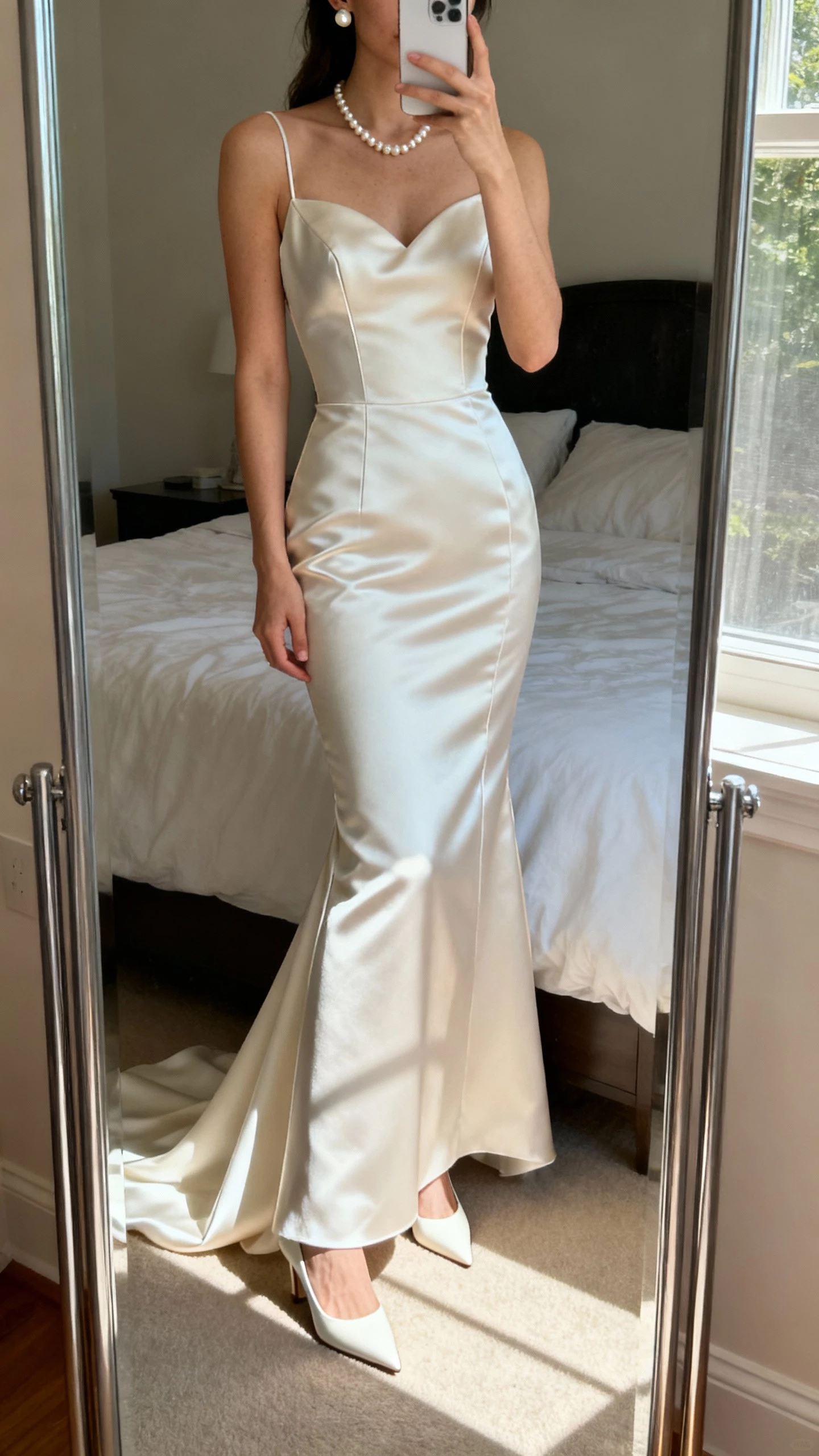 iPhone mirror selfie of a woman wearing a sculpted satin mermaid wedding dress with clean seams and a minimalist train, simple pearl studs and sleek pointed bridal heels, face not visible, cropped at neck, bedroom mirror with natural window light, natural iPhone photo quality