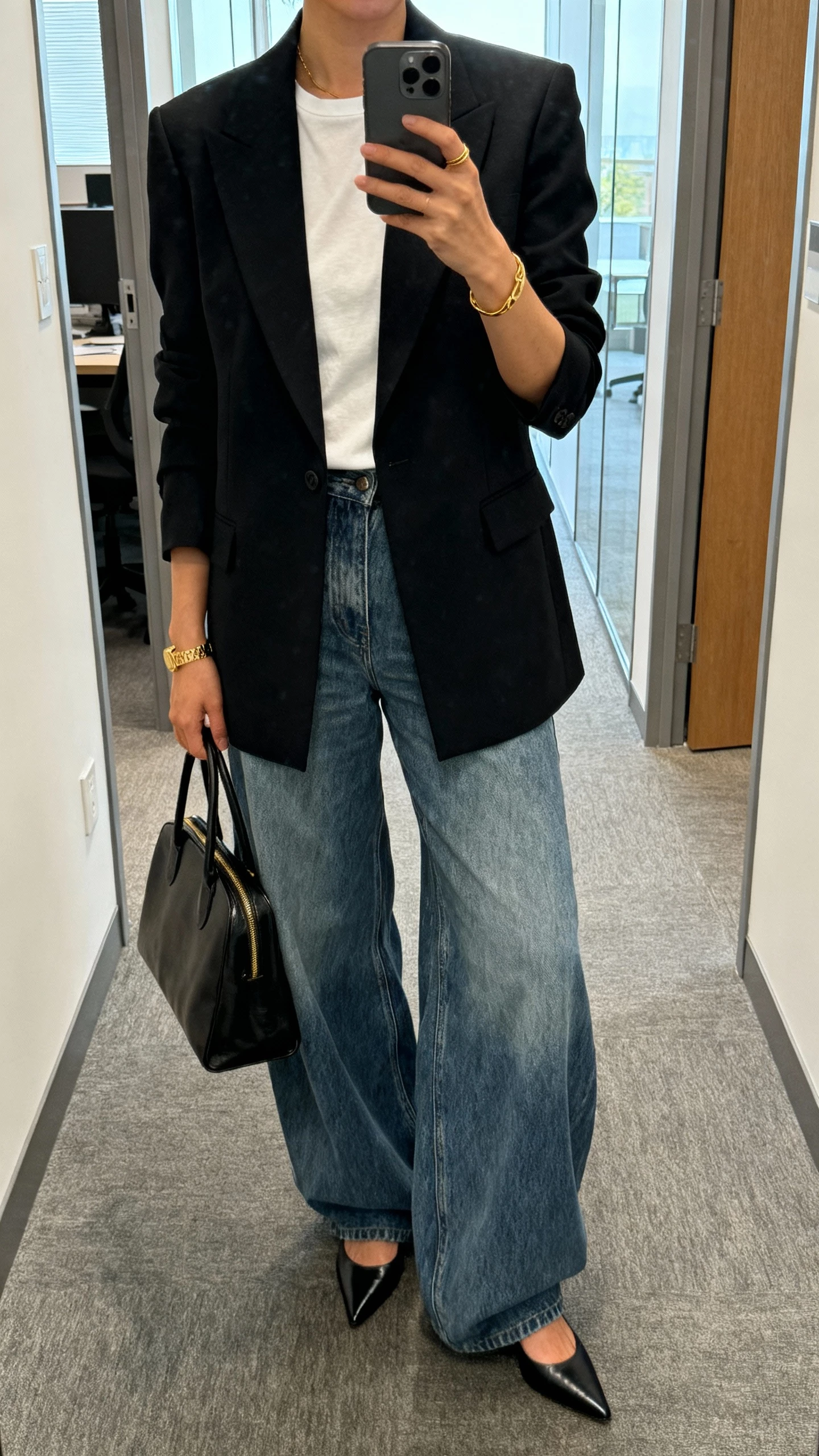 iPhone mirror selfie of a woman wearing a sharp power blazer over a simple tee with puddle baggy jeans, pointed heels, structured tote, minimal gold jewelry; office hallway mirror, face not visible, cropped at neck, natural morning light, natural iPhone photo quality.