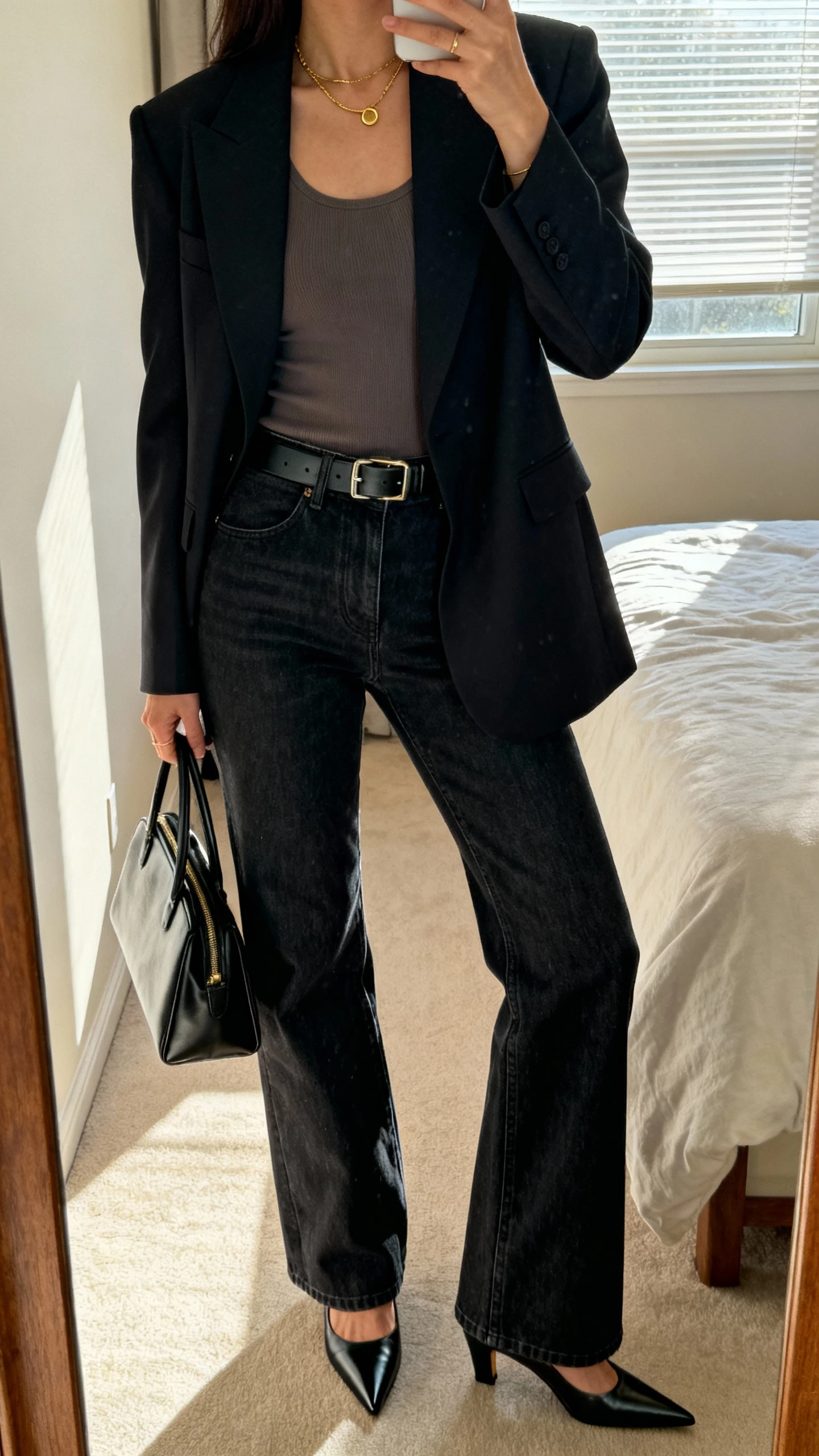 iPhone mirror selfie of a woman wearing a sharp tailored blazer over a fitted top, dark-wash bootcut jeans, sleek belt, pointed-toe heels, structured handbag, minimal gold jewelry; face not visible, cropped at neck, bedroom mirror with morning window light, natural iPhone photo quality.