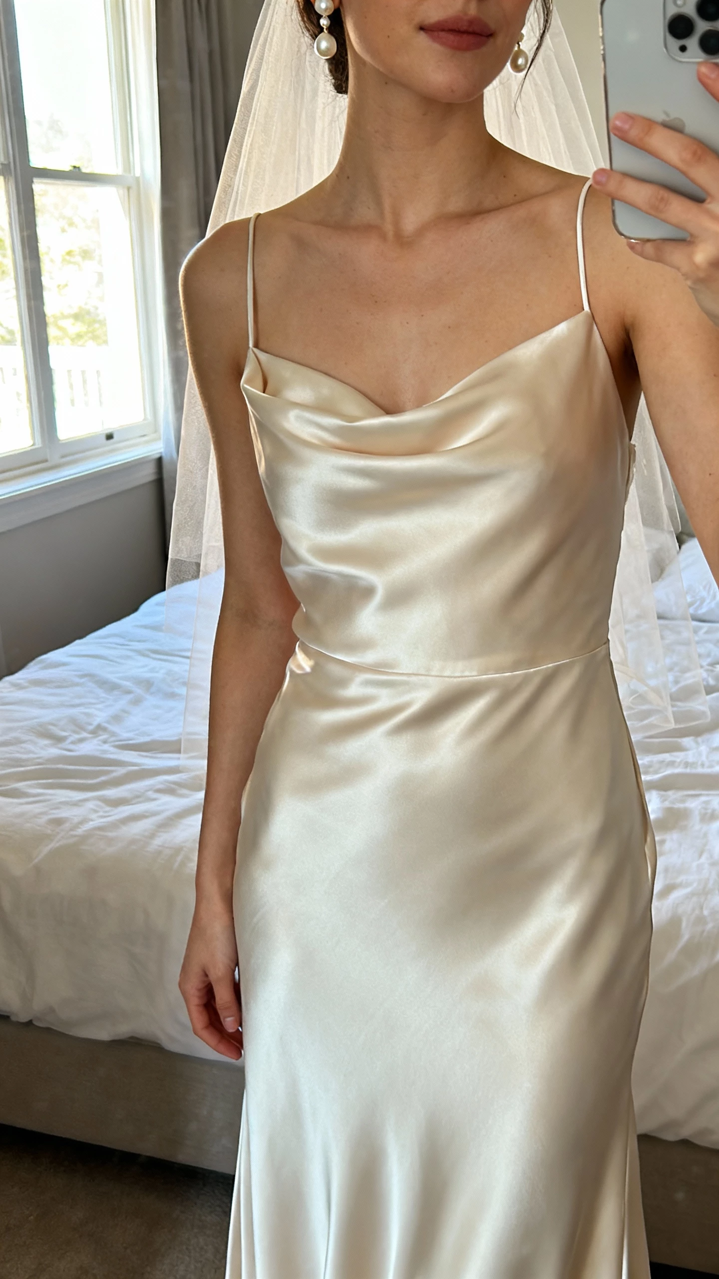 iPhone mirror selfie of a woman wearing a silk satin wedding gown with liquid-like drape, bias-cut slip silhouette, delicate spaghetti straps, simple veil, pearl drop earrings, face not visible, cropped at neck, bedroom mirror with morning window light, natural iPhone photo quality