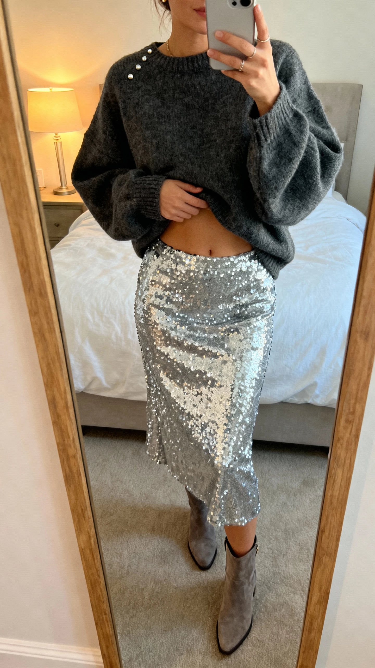 iPhone mirror selfie of a woman wearing a silver sequin skirt with an oversized charcoal sweater half-tucked, ankle boots, and simple studs, face not visible, bedroom mirror with soft lamp light, natural iPhone photo quality
