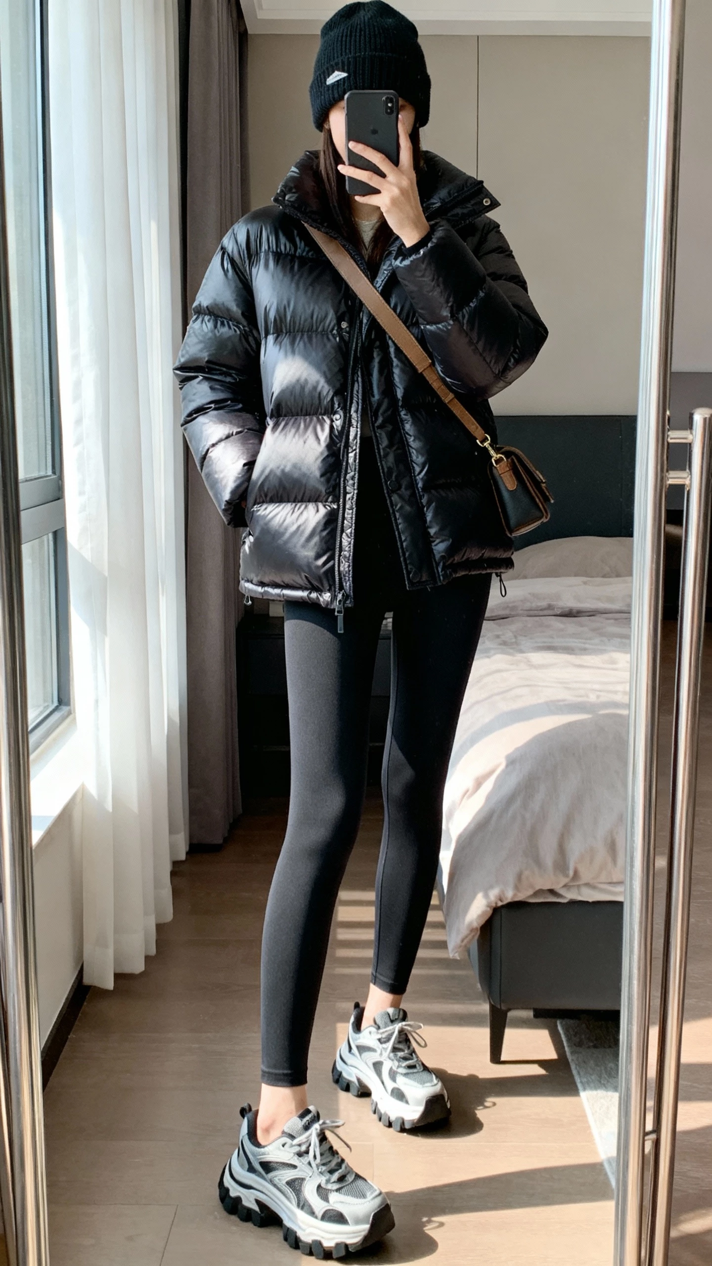 iPhone mirror selfie of a woman wearing a sleek puffer jacket, tailored leggings, chunky-soled sneakers, beanie, and crossbody bag; face not visible, cropped at neck, bedroom full-length mirror with morning window light, natural iPhone photo quality.