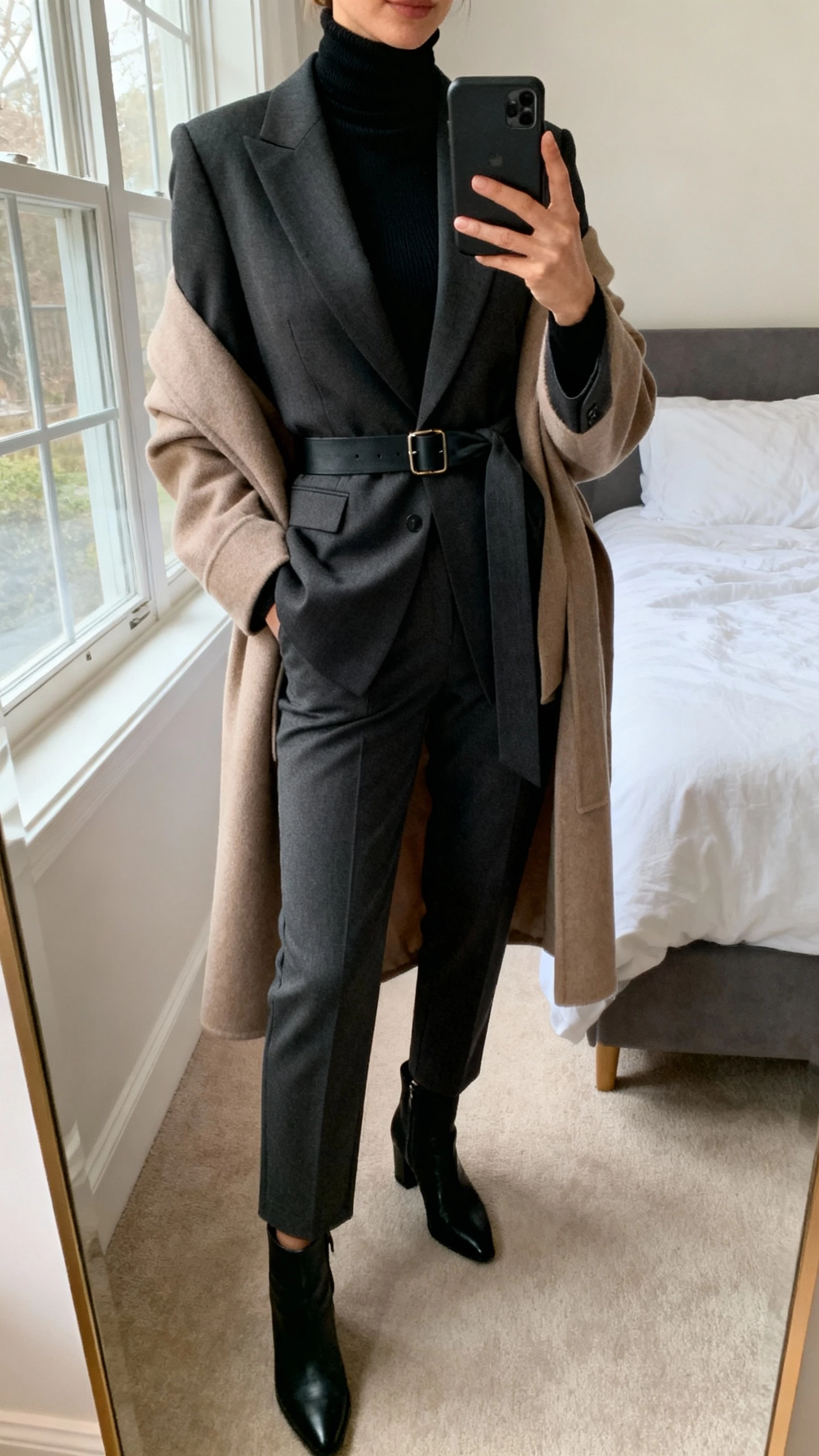iPhone mirror selfie of a woman wearing a slim black turtleneck, tailored blazer cinched with a belt, and a wrap coat layered on top, with tailored trousers and ankle boots, face not visible, cropped at neck, bedroom mirror, natural window light, natural iPhone photo quality.