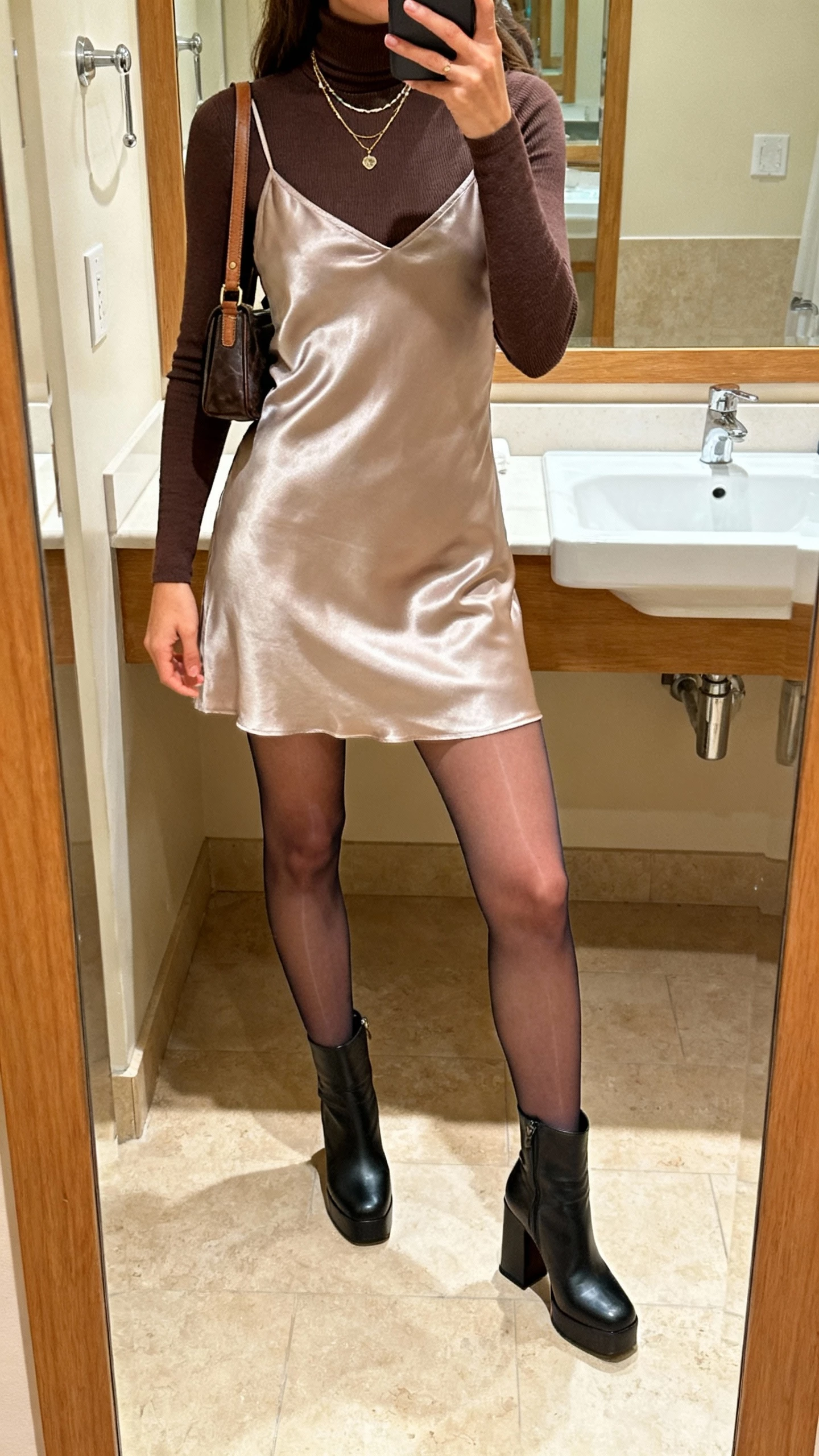 iPhone mirror selfie of a woman wearing a slim turtleneck under a satin slip dress, sheer tights, block-heel boots, small shoulder bag, and layered necklaces, face not visible, bathroom mirror, warm indoor lighting, natural iPhone photo quality.