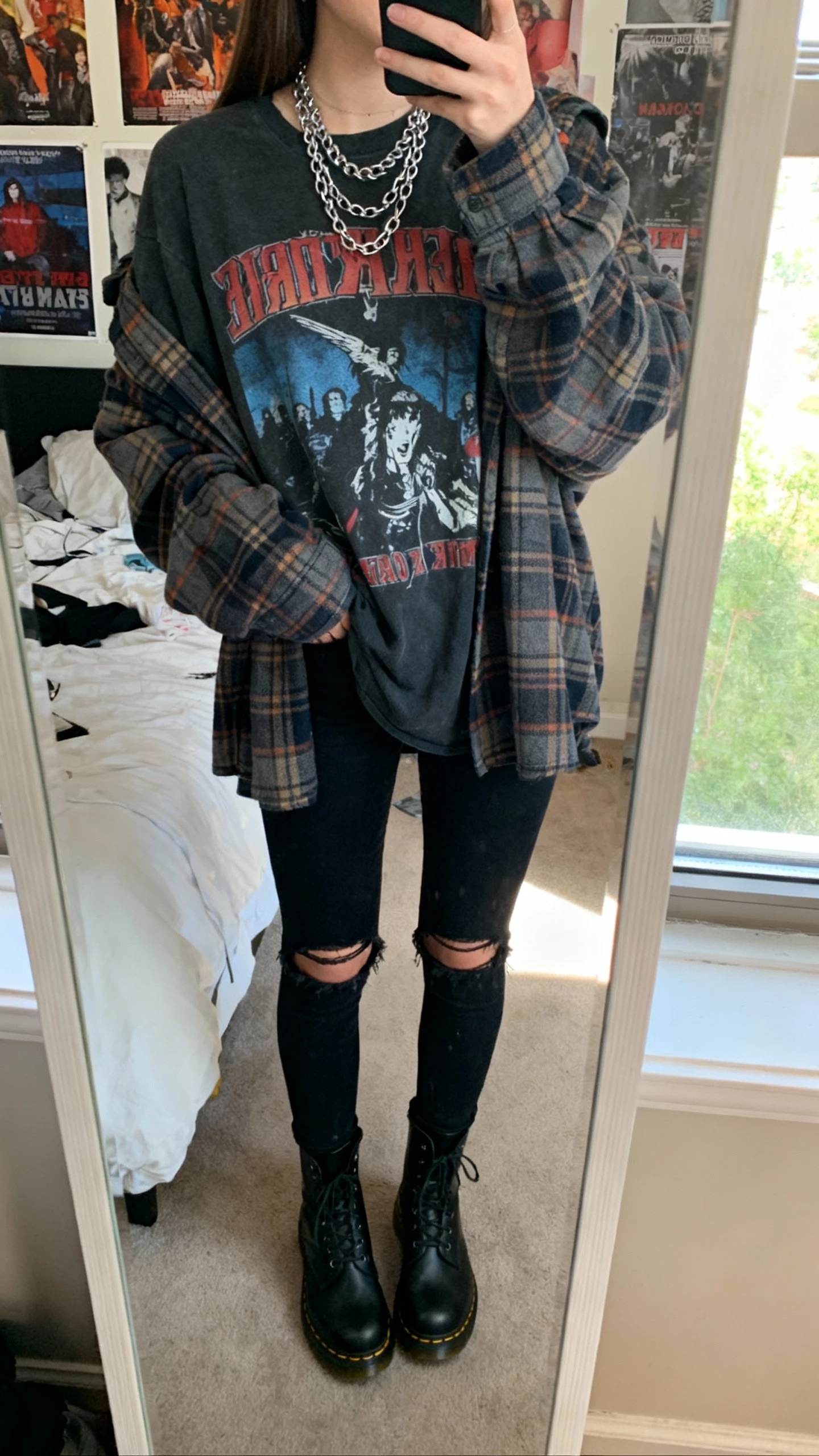 iPhone mirror selfie of a woman wearing a slouchy plaid flannel over a vintage band tee, black ripped skinny jeans, black Doc Martens, layered silver chains, face not visible, cropped at neck, messy bedroom mirror with posters, natural window light, natural iPhone photo quality.