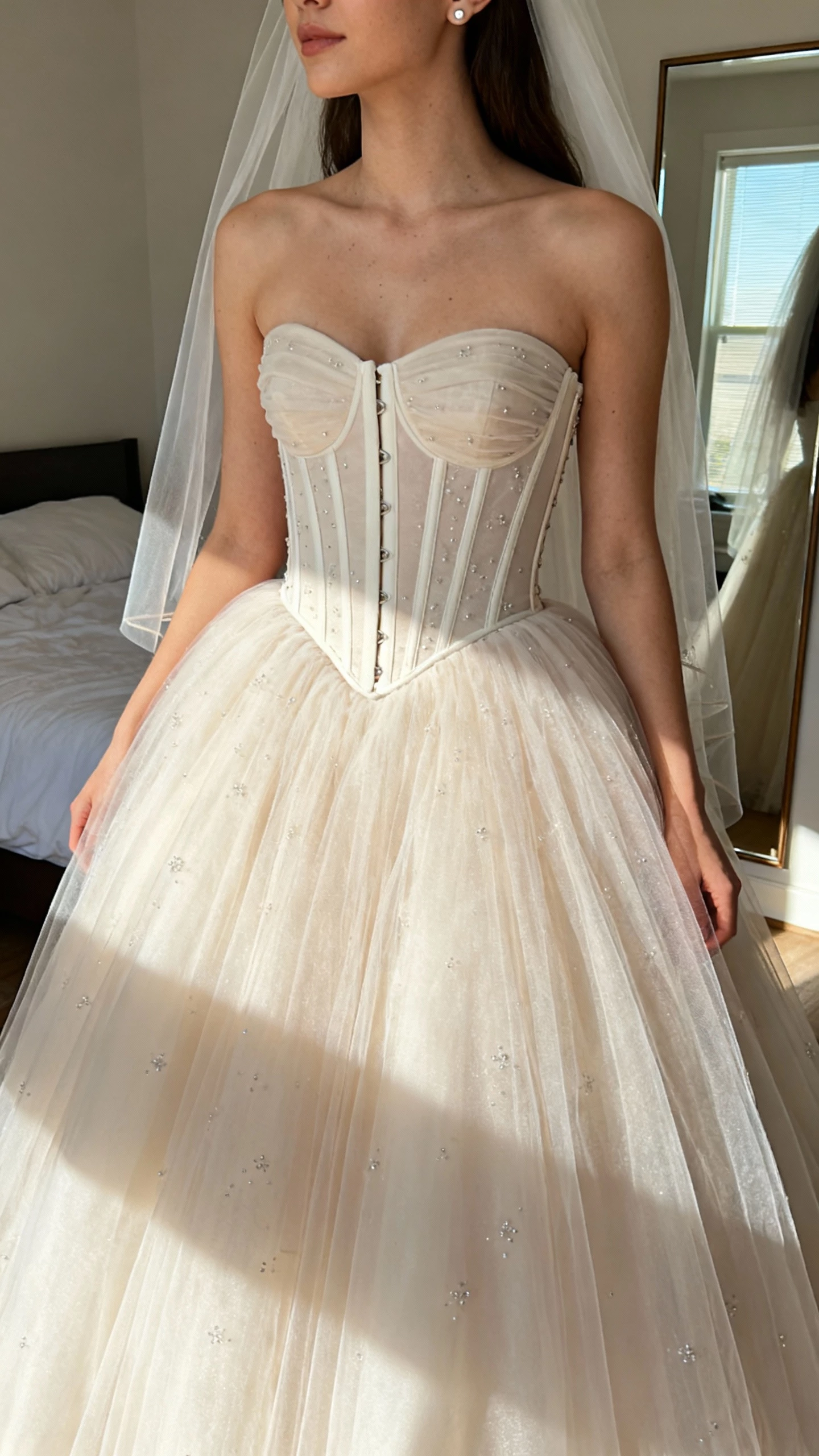 iPhone mirror selfie of a woman wearing a strapless corseted ballgown with a structured bodice, full tulle skirt, subtle beading, long veil, and simple stud earrings, face not visible, cropped at neck, bedroom mirror with natural window light, natural iPhone photo quality.