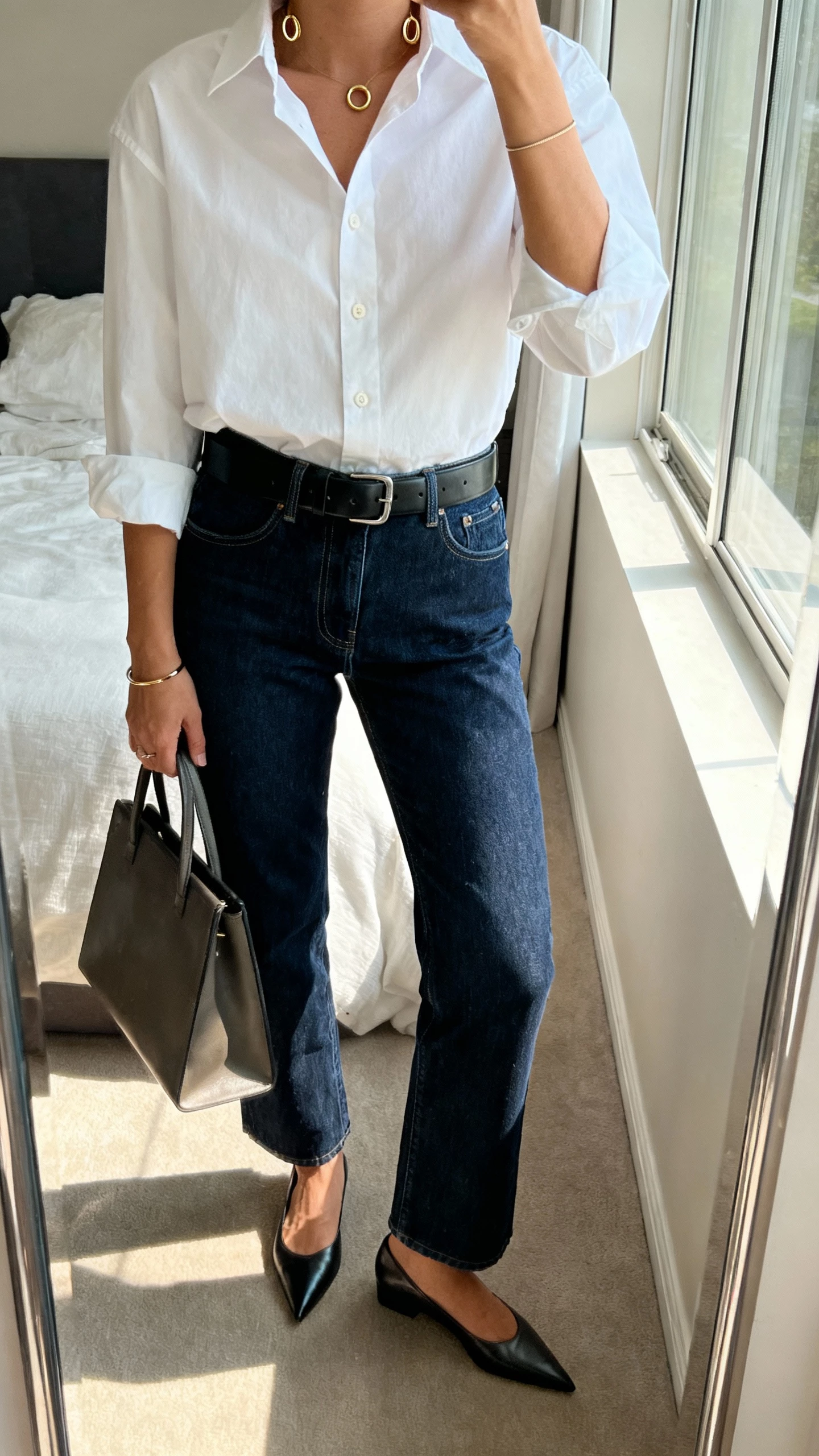 iPhone mirror selfie of a woman wearing a structured white button-up shirt tucked into dark blue straight-leg jeans with a black leather belt, pointed-toe flats, minimal gold huggie hoops and thin bracelet, carrying a structured tote; cropped at neck, bedroom mirror, natural window light, face not visible, natural iPhone photo quality.