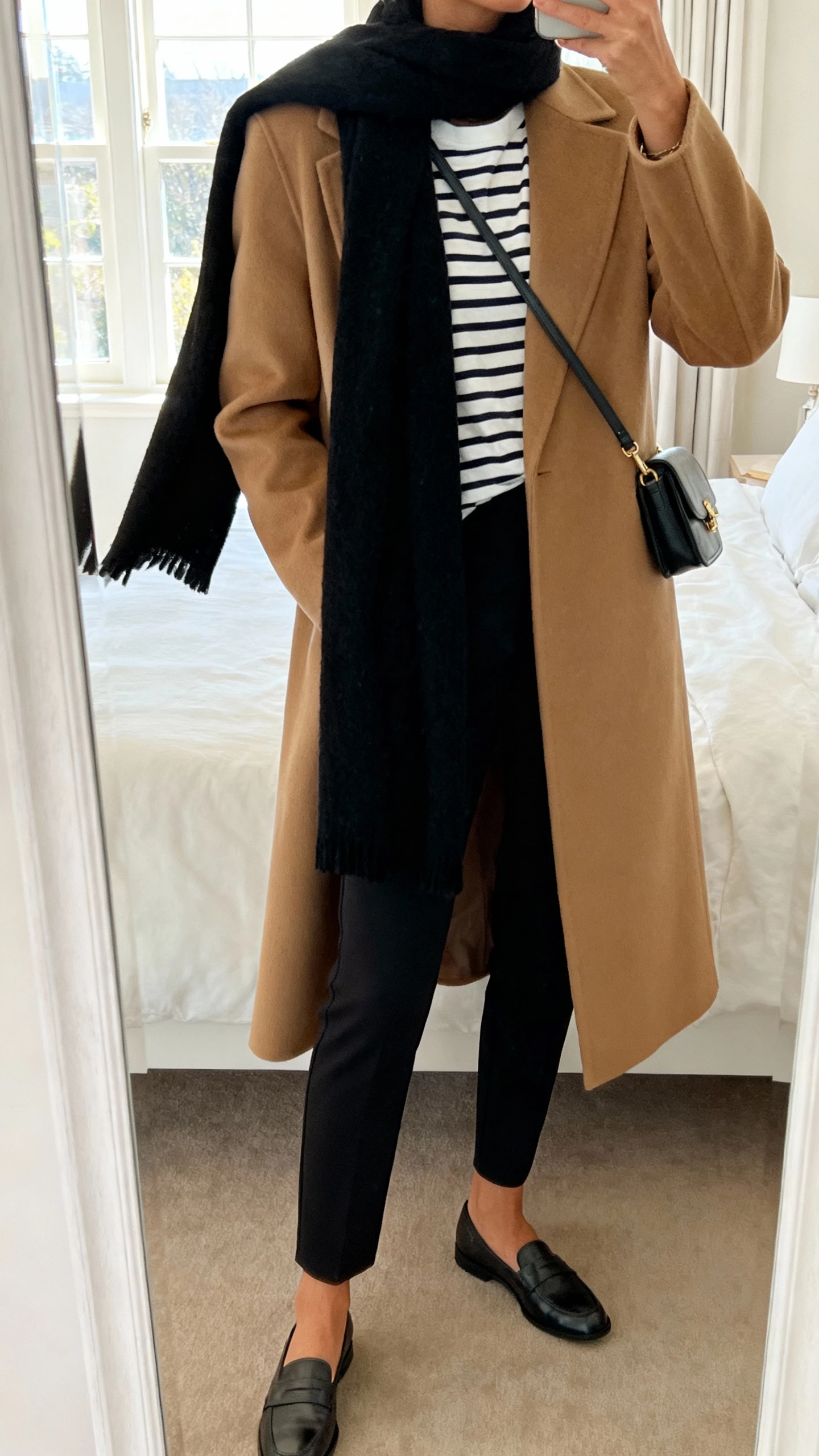 iPhone mirror selfie of a woman wearing a tailored camel coat over a striped Breton tee with slim black trousers, draping a long black wool scarf Parisian-style with one tail tossed over the shoulder, loafers and a small crossbody bag, face not visible, cropped at neck, bedroom mirror, soft morning window light, natural iPhone photo quality.