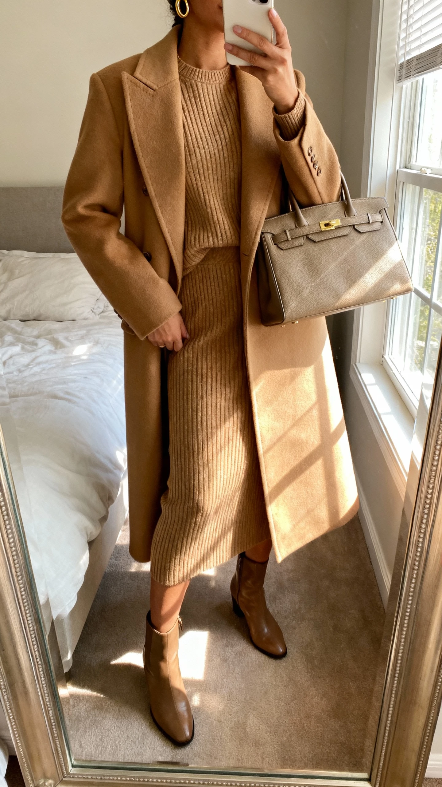 iPhone mirror selfie of a woman wearing a tailored camel coat over a matching ribbed knit set (crewneck sweater and knit midi skirt), sleek ankle boots, structured tote, simple gold hoops, face not visible, cropped at neck, bedroom mirror with natural window light, natural iPhone photo quality.