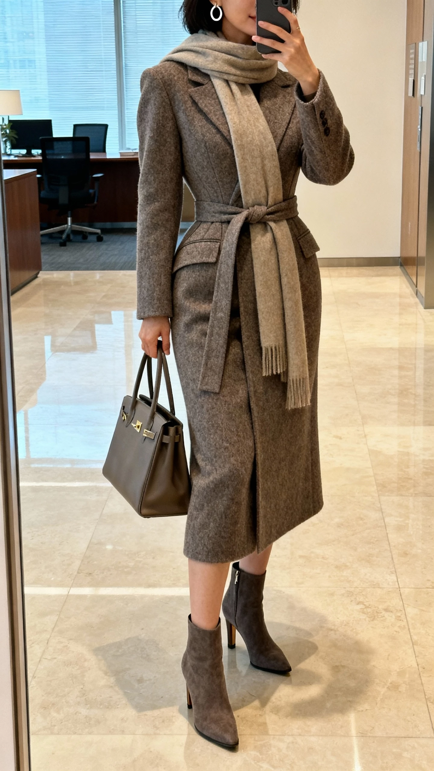 iPhone mirror selfie of a woman wearing a tailored wool coat with a long scarf belted at the waist to create an hourglass shape, heeled ankle boots, structured handbag, simple hoop earrings, face not visible, cropped at shoulders, office lobby mirror, natural indoor lighting, iPhone photo quality.