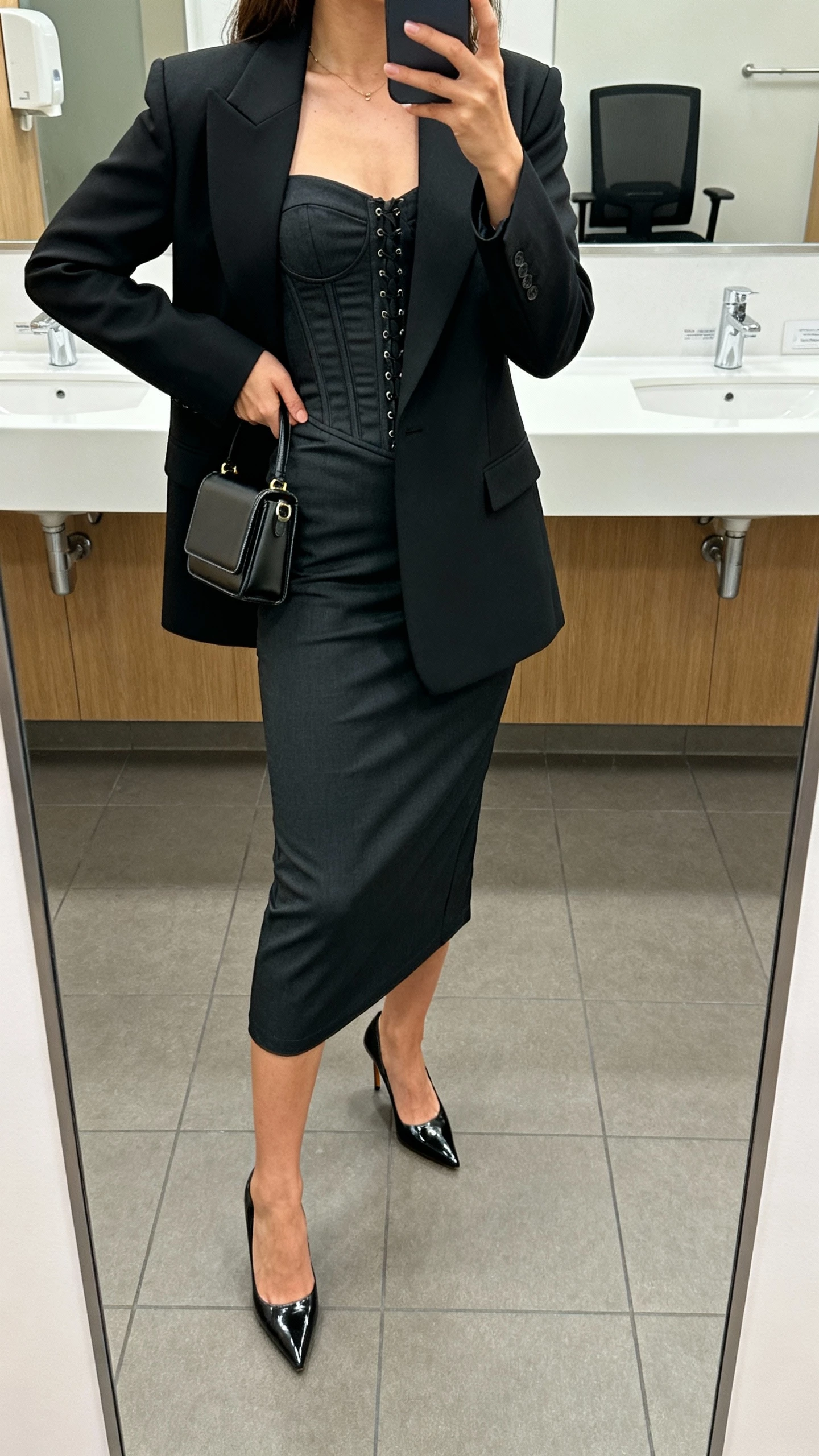 iPhone mirror selfie of a woman wearing a tailored corset midi dress under a sharp power blazer with sleek pumps and a structured mini bag, face not visible, cropped at neck, office restroom mirror, natural indoor light, iPhone photo quality.