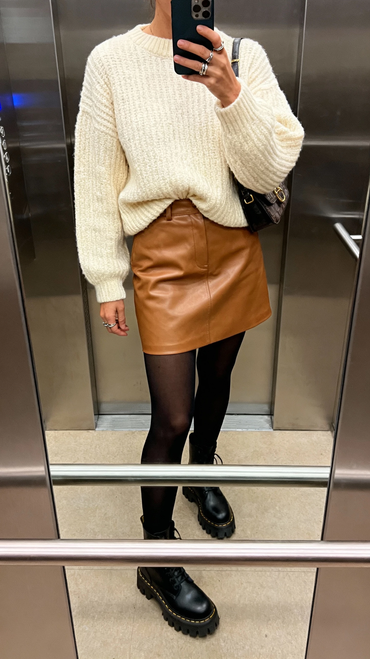 iPhone mirror selfie of a woman wearing a tan leather mini skirt with a chunky cream sweater half-tucked, black tights, lug-sole ankle boots, small shoulder bag, layered rings, face not visible, cropped at neck, elevator mirror, ambient evening light, casual iPhone photo.