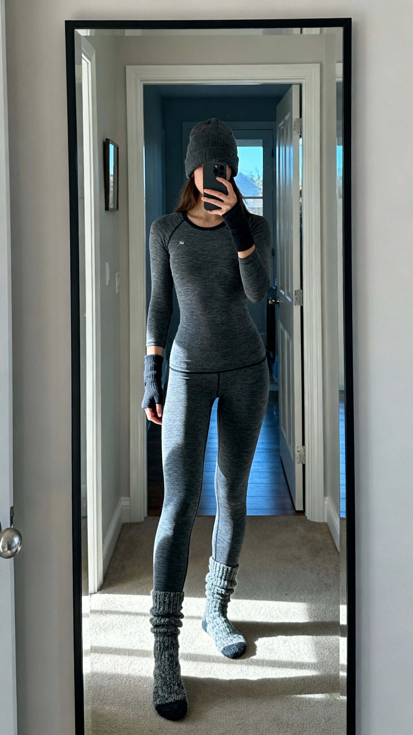 iPhone mirror selfie of a woman wearing a thermal two-piece with thumbholes (slim thermal top and matching leggings) plus wool socks and a beanie, face not visible, entryway mirror with cool morning light, natural iPhone photo.