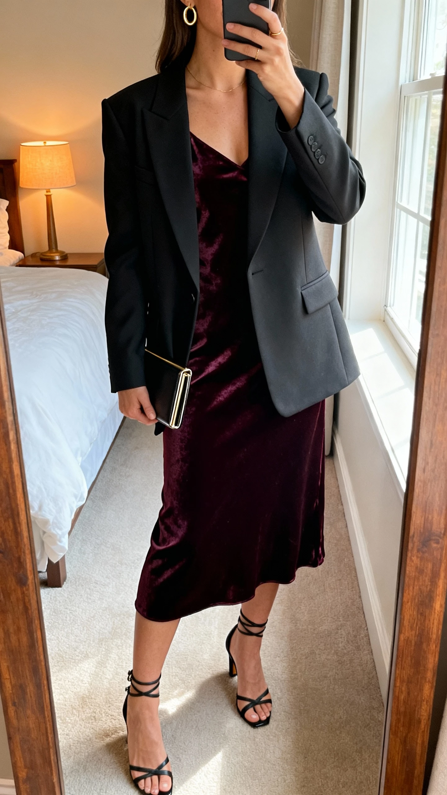 iPhone mirror selfie of a woman wearing a velvet slip dress under a sharp power blazer, strappy heels, delicate gold hoop earrings and a slim clutch, face not visible, cropped at neck, bedroom full-length mirror with warm lamp and window light, natural iPhone photo quality.