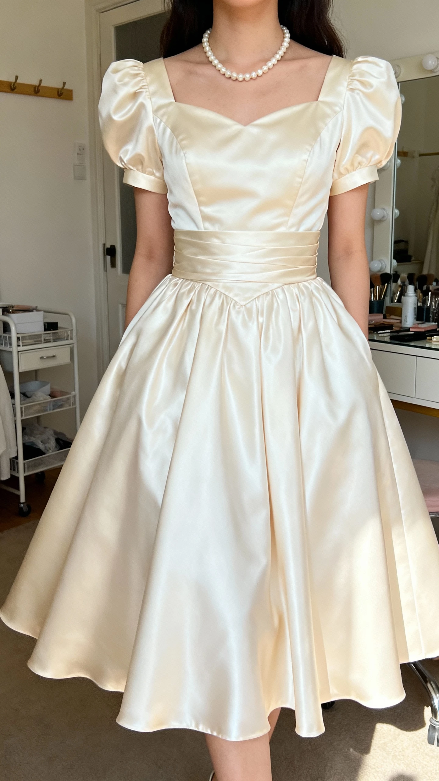 iPhone mirror selfie of a woman wearing a vintage-inspired satin tea-length bridal dress with strong waist emphasis and swingy skirt, classic pearls, face not visible, retro-styled dressing room, natural indoor light, natural iPhone photo quality