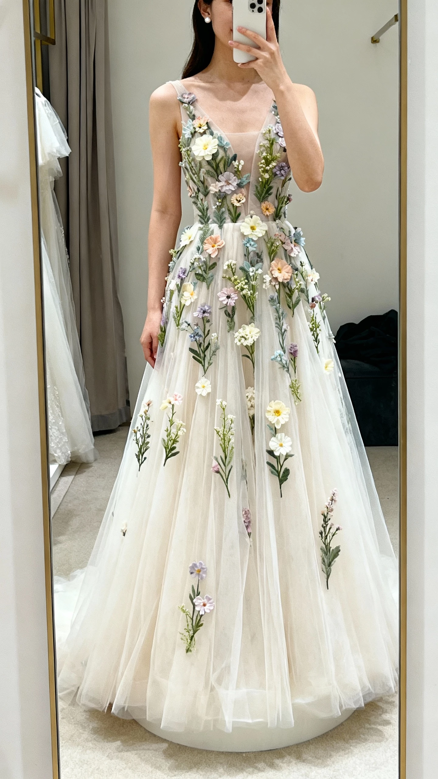 iPhone mirror selfie of a woman wearing a whimsical tulle A-line wedding gown adorned with 3D wildflower appliqués, soft tulle skirt, simple stud earrings, face not visible, dressing room mirror, gentle indoor lighting, natural iPhone photo quality