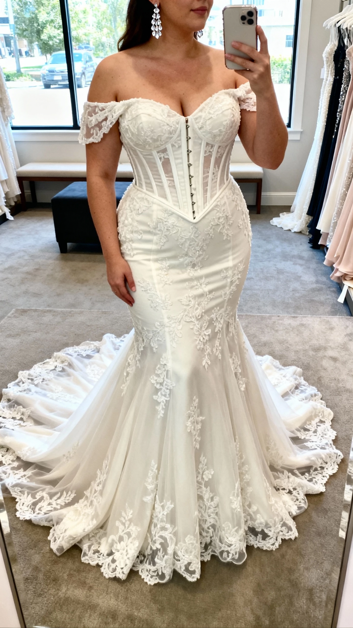 iPhone mirror selfie of a woman wearing an Allure Bridals Curve sculpted mermaid wedding gown with corset bodice, off-the-shoulder sleeves, lace appliqués, and dramatic cathedral train, paired with crystal drop earrings; face not visible, cropped at neck, boutique fitting room, natural window light, natural iPhone photo quality.