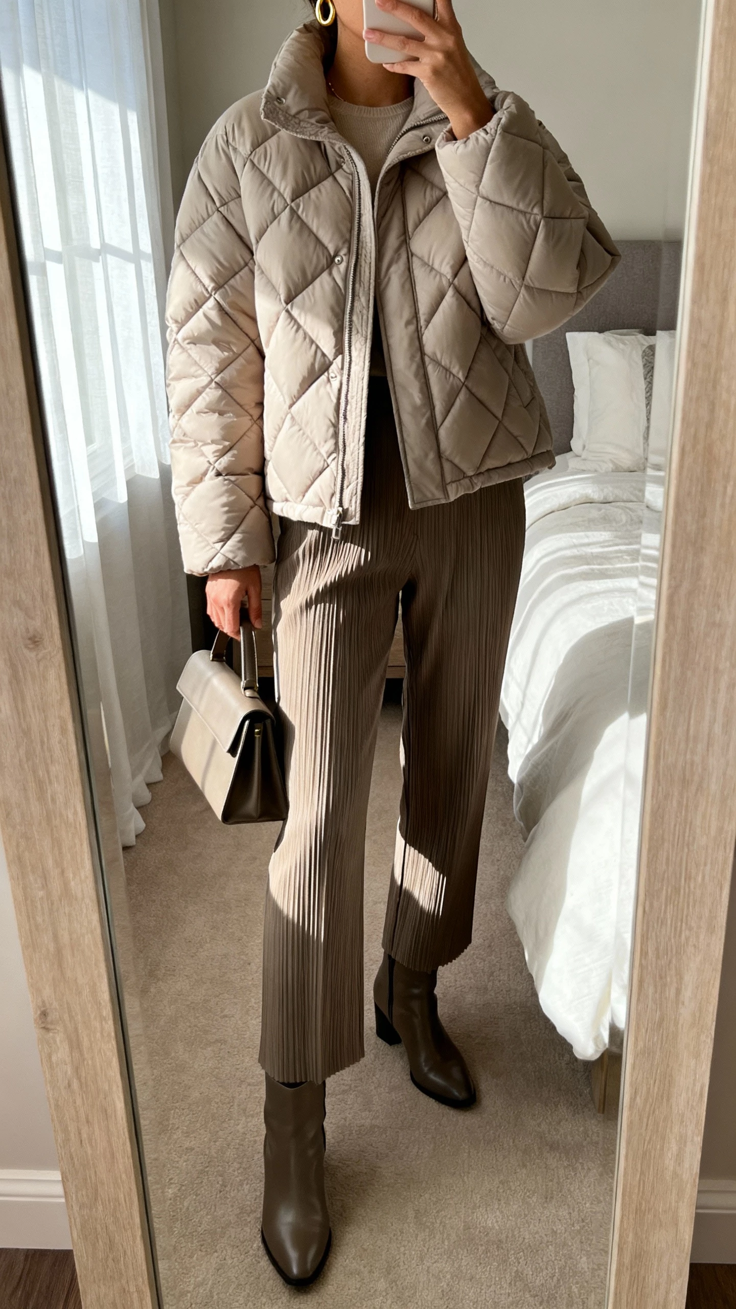 iPhone mirror selfie of a woman wearing an elevated quilted puffer jacket, tailored pleated trousers, sleek leather ankle boots, minimal gold hoops, structured tote, face not visible, cropped at neck, bedroom full-length mirror, soft morning window light, natural iPhone photo quality.