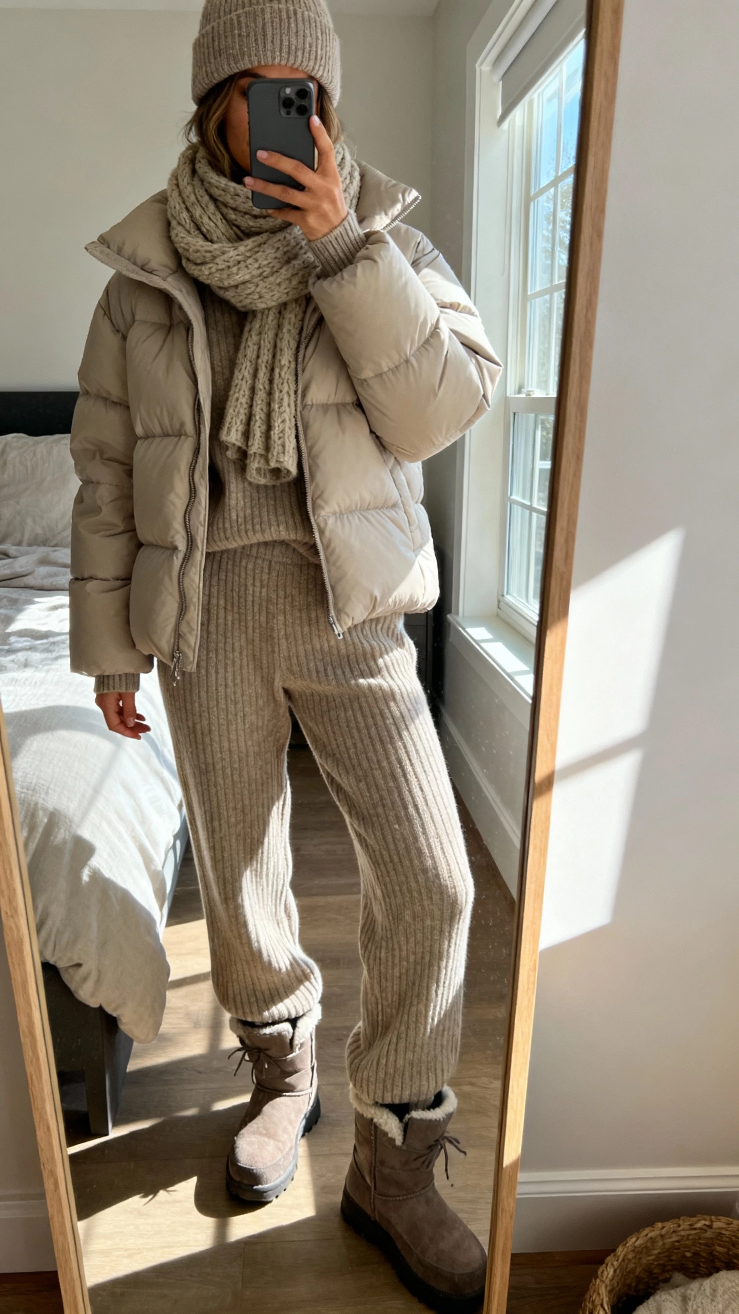 iPhone mirror selfie of a woman wearing an elevated puffer jacket over a matching ribbed knit set (sweater and knit pants), chunky scarf, wool beanie, and winter boots, face not visible, cropped at neck, bedroom mirror with natural window light, natural iPhone photo quality.