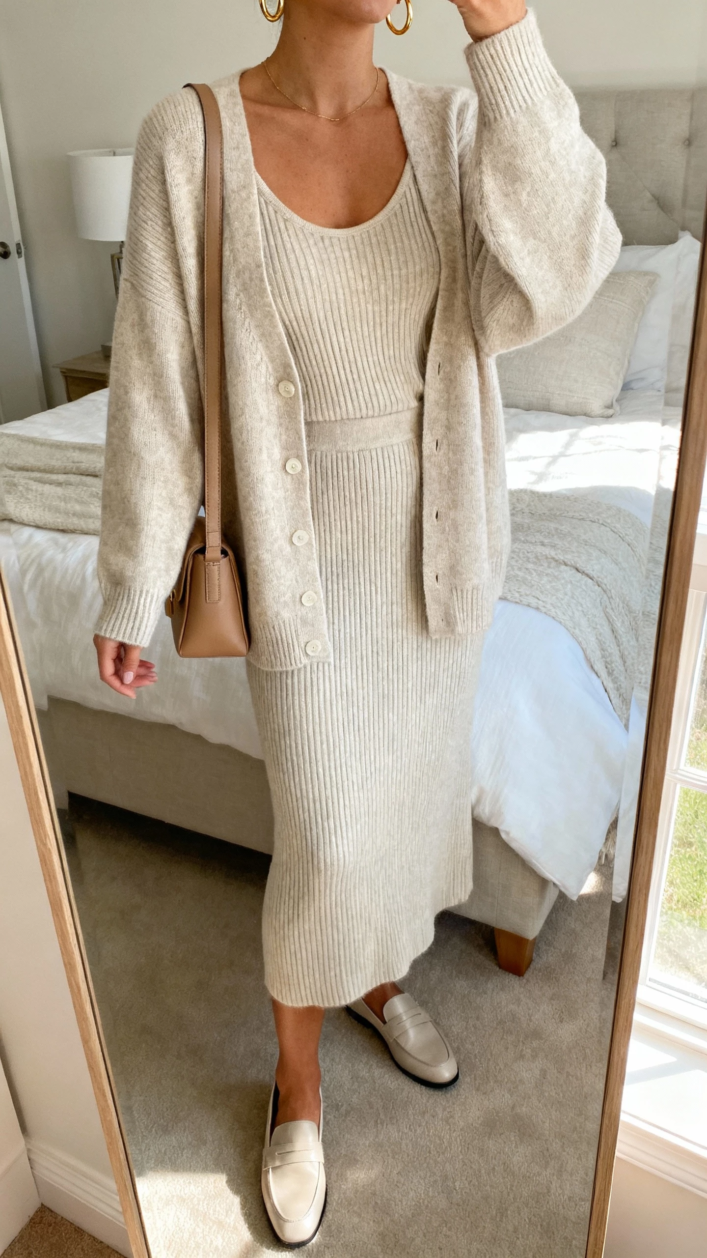 iPhone mirror selfie of a woman wearing an elevated knit sweater set: matching fine-gauge cashmere cardigan and tank with coordinating ribbed midi skirt, delicate gold hoops, minimalist leather shoulder bag, sleek loafers, face not visible, cropped at neck, bedroom mirror with morning window light, natural iPhone photo quality