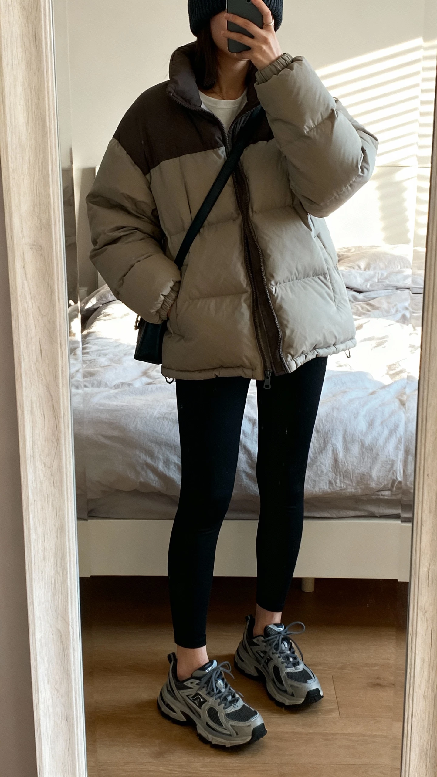 iPhone mirror selfie of a woman wearing an elevated puffer jacket, black leggings, chunky sneakers, beanie, crossbody bag, face not visible, cropped at neck, bedroom mirror with natural morning light, natural iPhone photo quality.