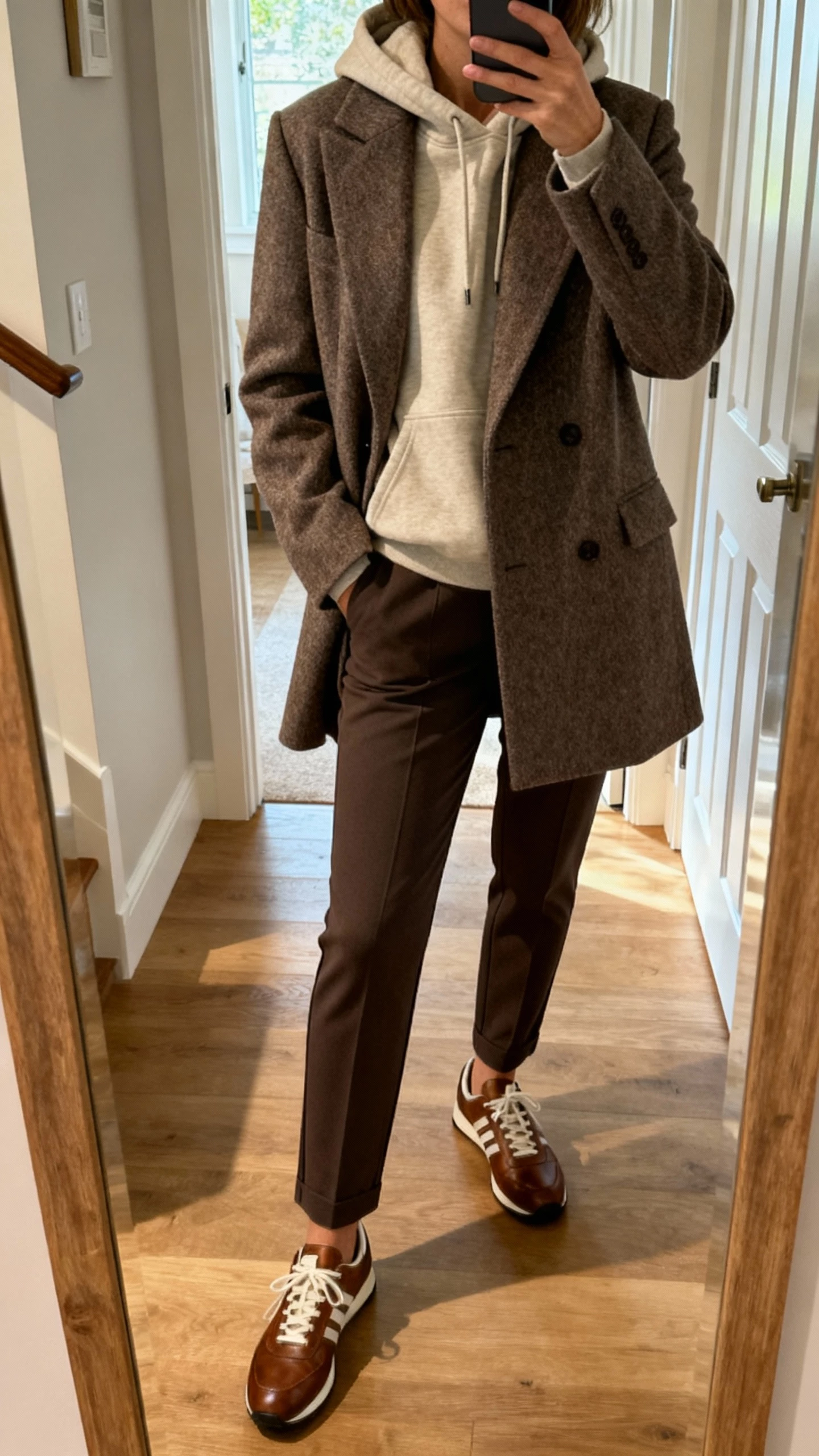 iPhone mirror selfie of a woman wearing an elevated hoodie layered under a structured wool coat, tailored joggers or slim trousers, leather sneakers, face not visible, entryway mirror, morning light, natural iPhone photo quality.
