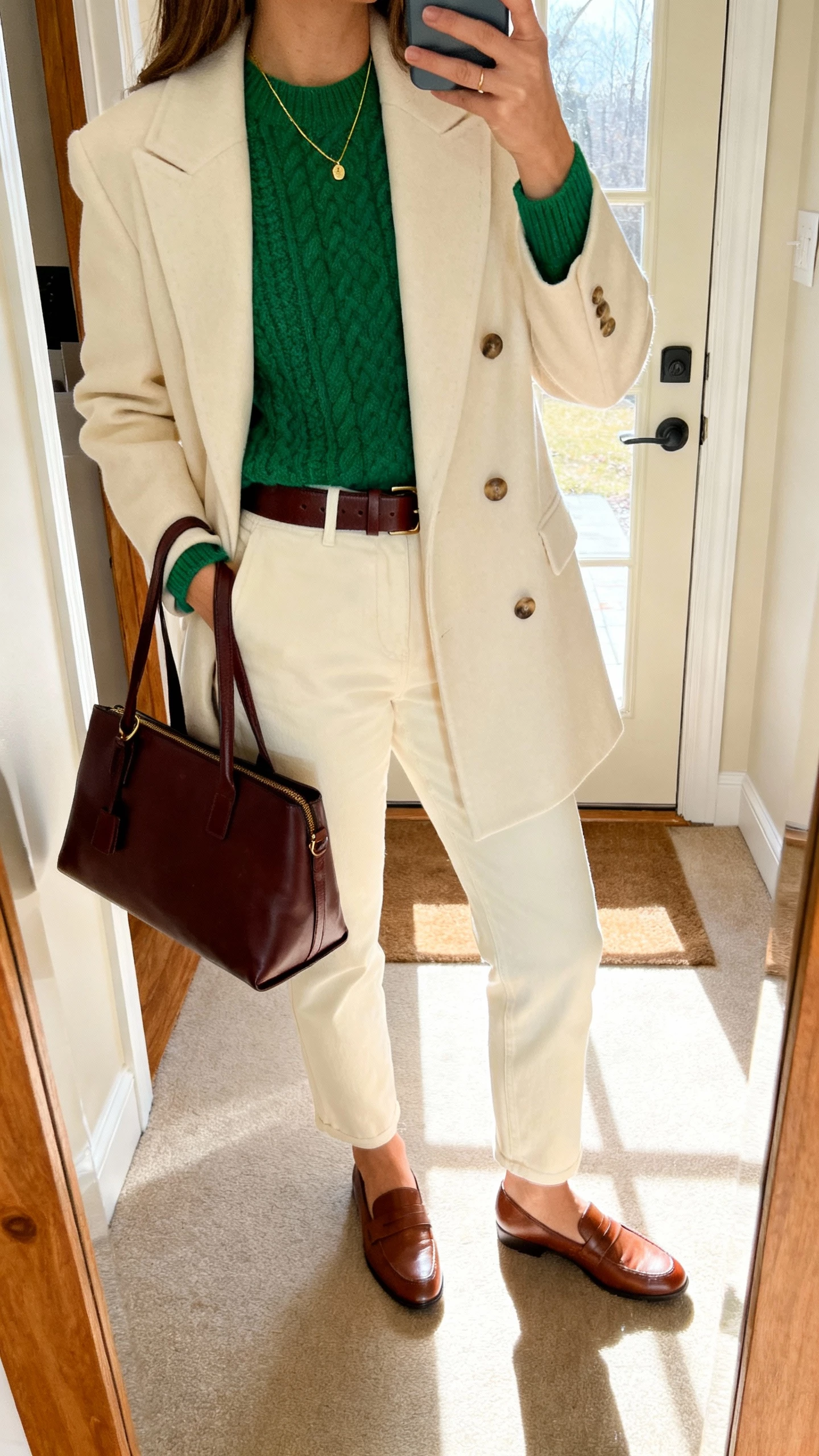 iPhone mirror selfie of a woman wearing an emerald knit sweater, cream tailored coat, espresso leather belt and tote, cream trousers, brown loafers, delicate gold necklace, face not visible, entryway mirror with daylight from door, natural iPhone photo quality