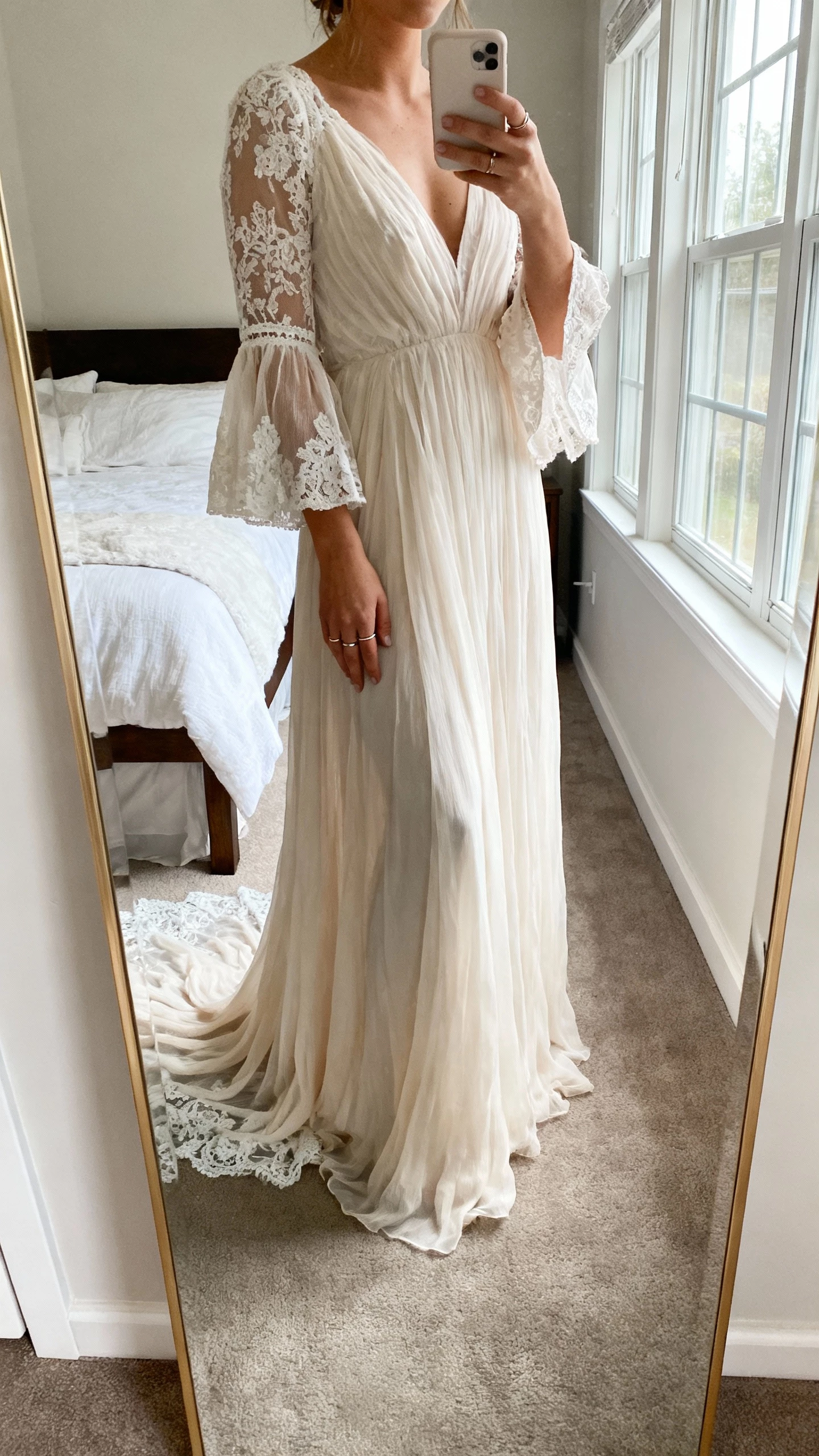 iPhone mirror selfie of a woman wearing an ethereal lace bell-sleeve boho wedding gown that drapes and moves fluidly, delicate lace train, minimal rings, face not visible, cropped at neck, bedroom full-length mirror, natural window light, natural iPhone photo quality