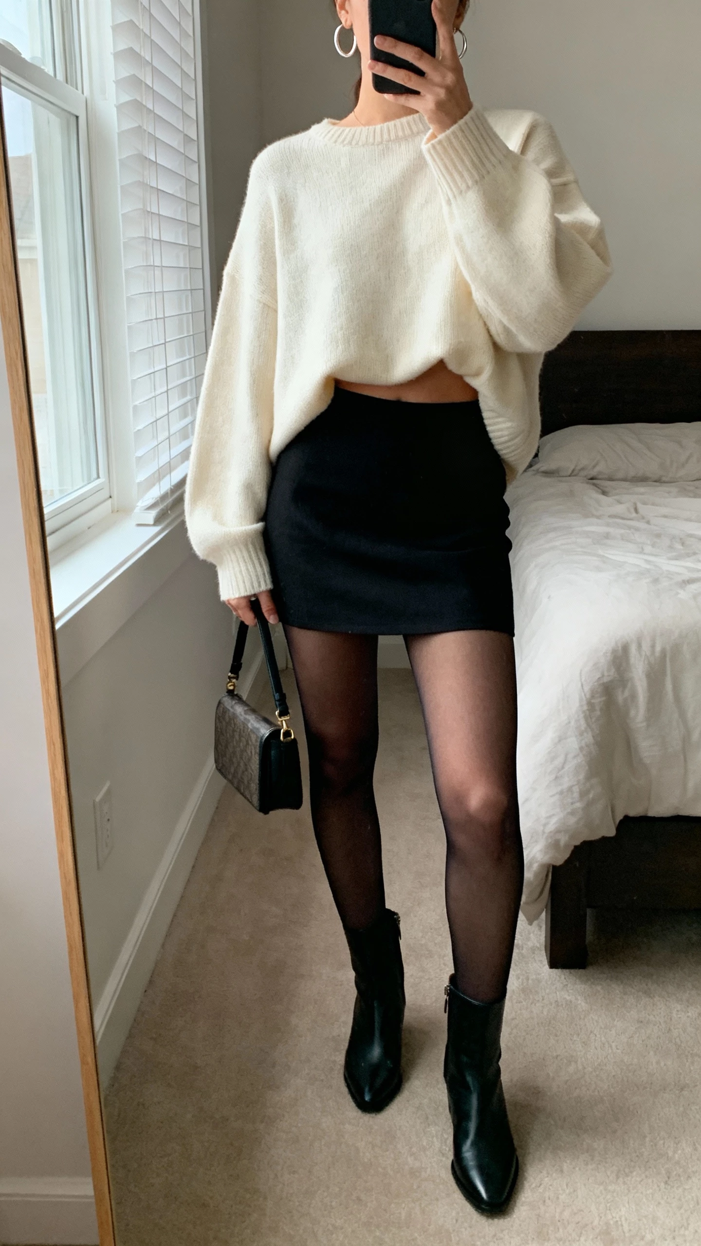 iPhone mirror selfie of a woman wearing an oversized cream sweater half-tucked into a black mini skirt, sheer tights, ankle boots, simple hoop earrings, small crossbody bag in hand, face not visible, cropped at neck, bedroom mirror with morning window light, natural iPhone photo quality.