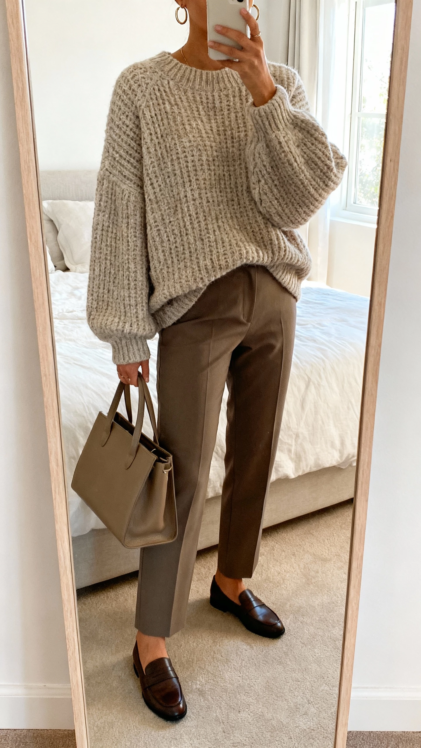 iPhone mirror selfie of a woman wearing an oversized fisherman sweater with tailored trousers and loafers, simple hoop earrings and a structured tote, face not visible, cropped at neck, bedroom full-length mirror, soft window light, natural iPhone photo quality