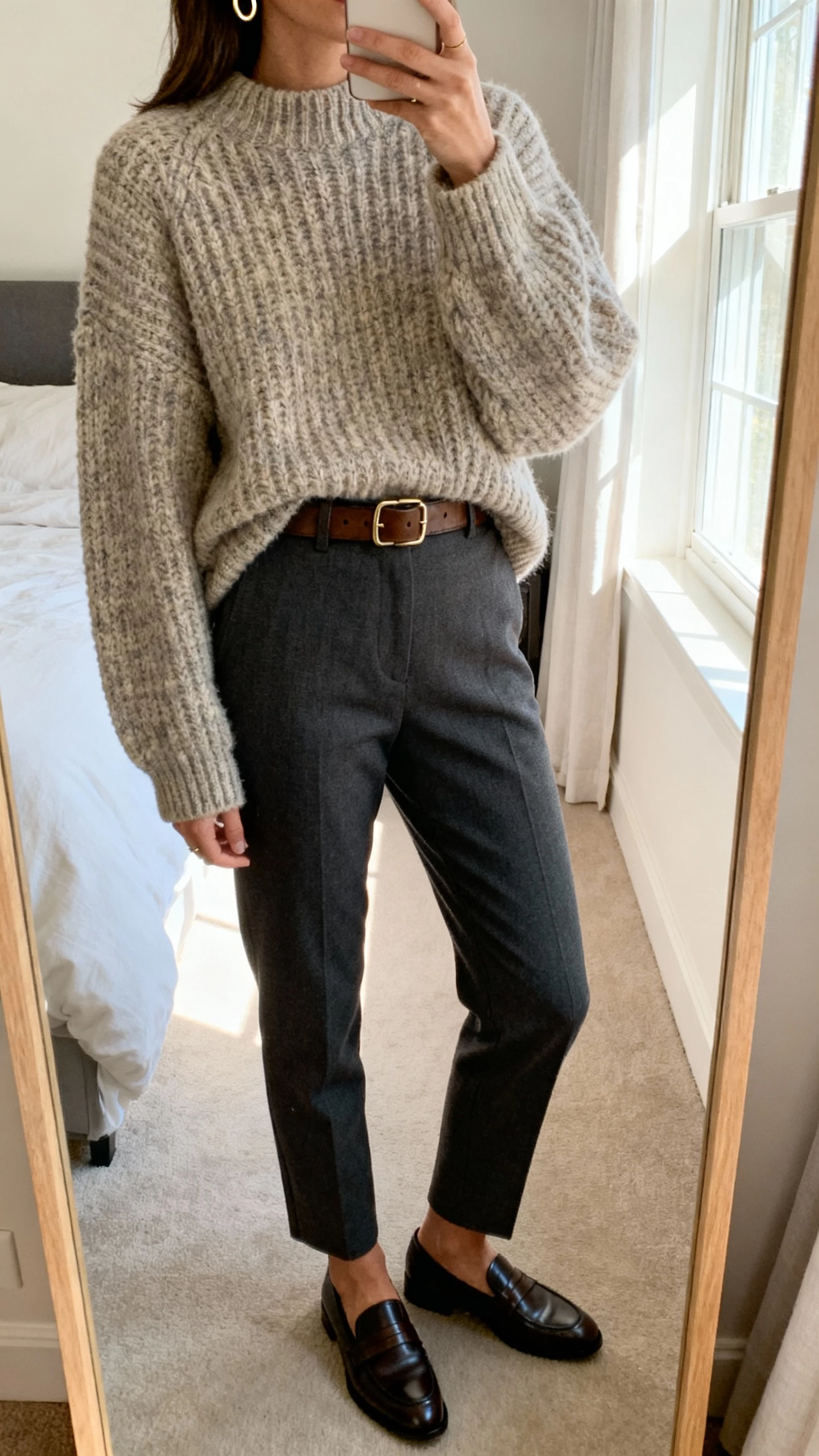 iPhone mirror selfie of a woman wearing an oversized chunky knit sweater, tailored trousers, sleek loafers, a leather belt, and minimal gold hoops, face not visible, cropped at neck, bedroom mirror with morning window light, natural iPhone photo quality.