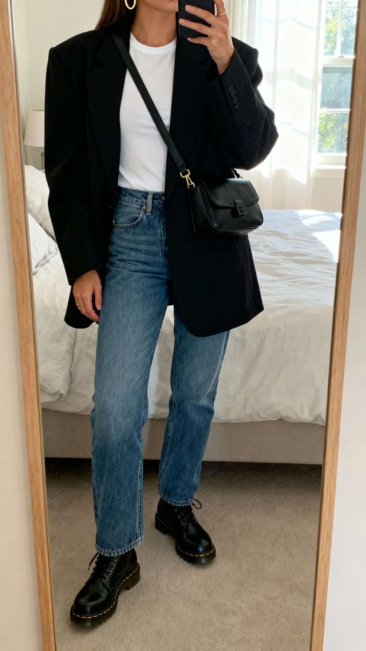iPhone mirror selfie of a woman wearing an oversized black blazer over a white fitted tee, straight-leg blue jeans, black Doc Martens, minimal gold hoops, and a black crossbody bag, face not visible, cropped at neck, bedroom mirror with morning window light, natural iPhone photo quality.