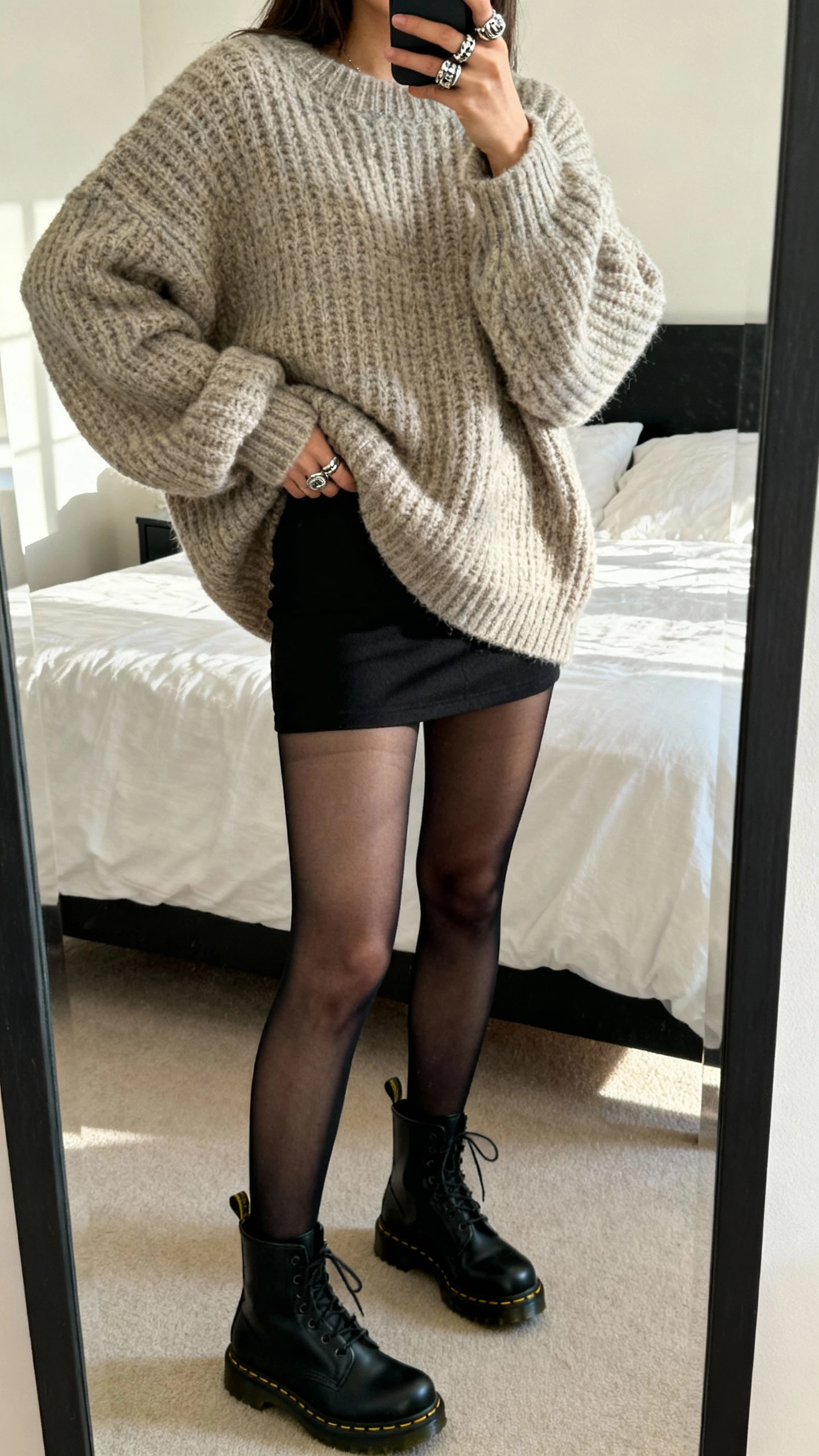 iPhone mirror selfie of a woman wearing an oversized chunky knit sweater with a black micro skirt, sheer black tights, black Doc Martens, chunky rings, face not visible, bedroom full-length mirror, morning light, natural iPhone photo quality.
