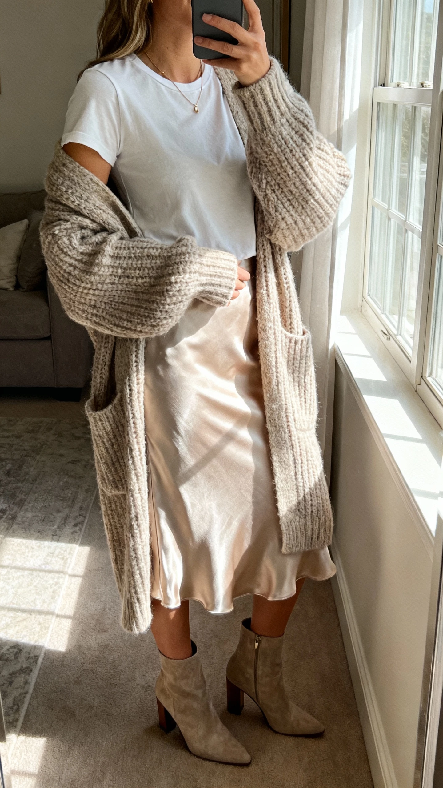 iPhone mirror selfie of a woman wearing an oversized chunky cardigan layered over a satin slip skirt, fitted tee, heeled ankle boots, delicate necklace, face not visible, living room mirror with window light, natural iPhone photo quality