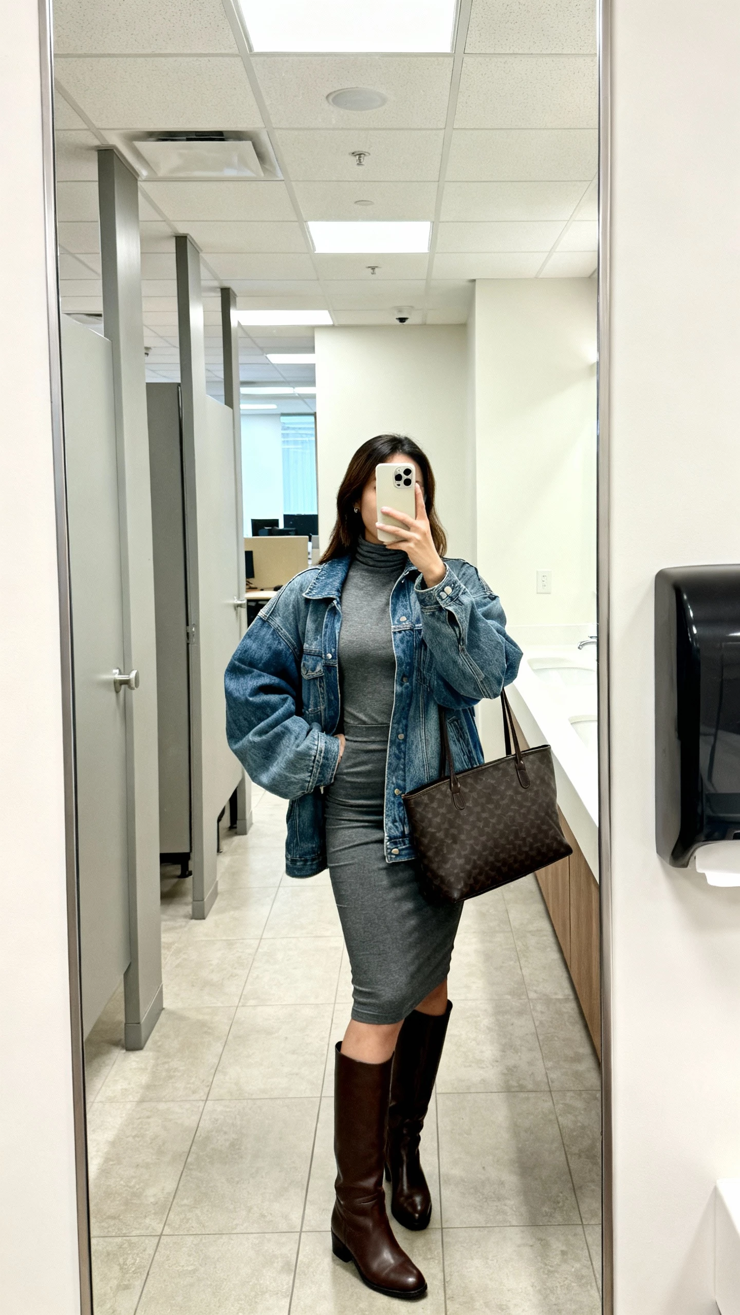 iPhone mirror selfie of a woman wearing an oversized jean jacket with a fitted turtleneck, pencil skirt, knee-high leather boots, structured tote, face not visible, office restroom mirror, bright office lighting, casual iPhone photo