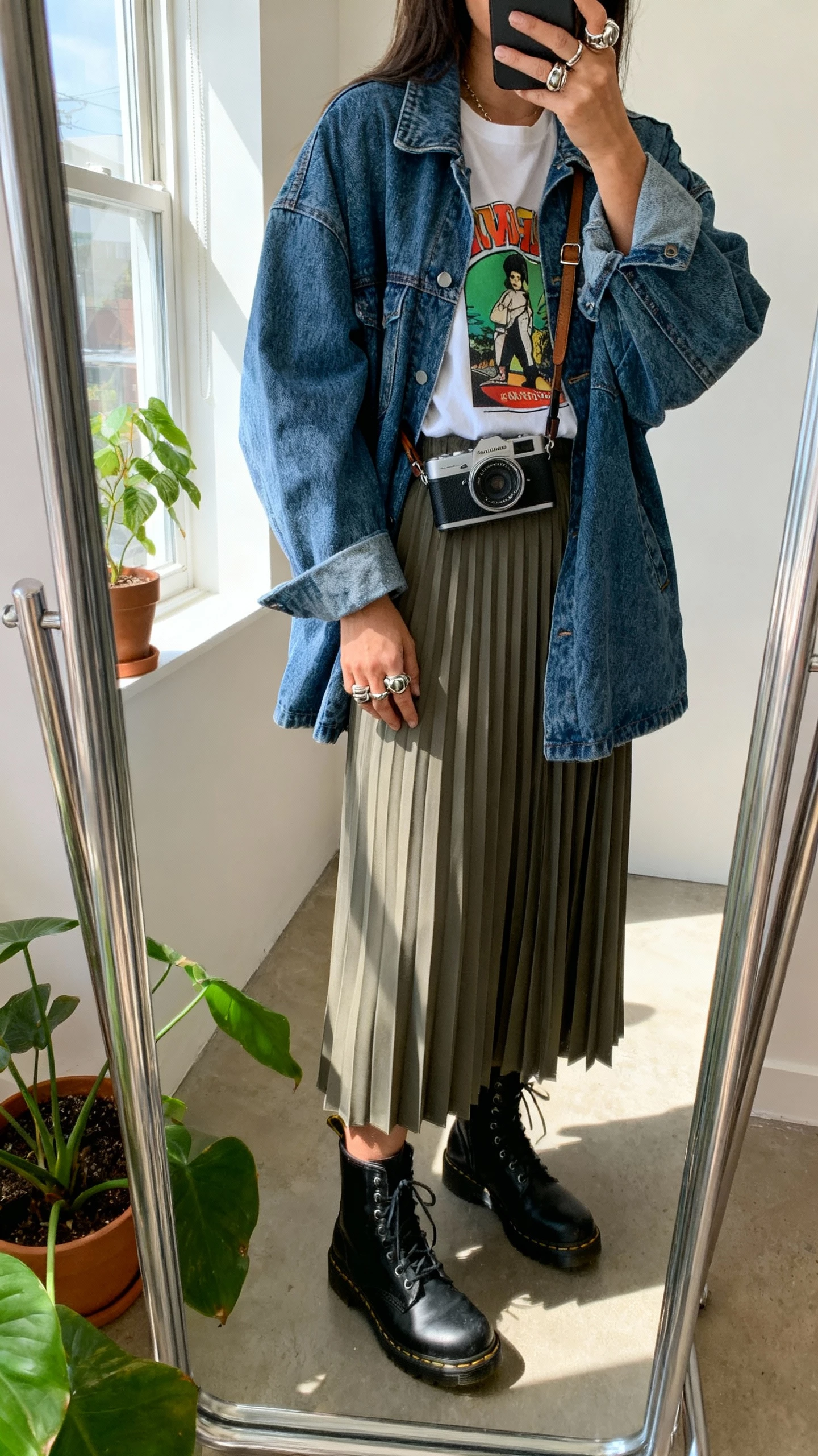 iPhone mirror selfie of a woman wearing an oversized jean jacket, graphic tee, pleated midi skirt, combat boots, chunky rings, crossbody camera bag, face not visible, studio corner mirror with plants, afternoon window light, casual iPhone photo