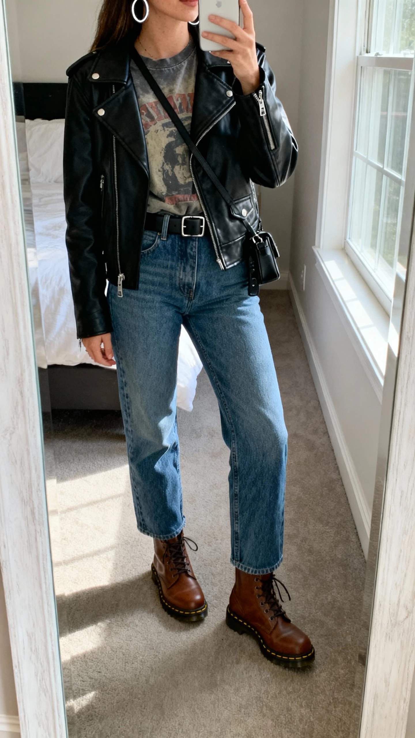 iPhone mirror selfie of a woman wearing brown Doc Martens with straight-leg blue denim, black leather moto jacket over a faded graphic tee, silver hoop earrings, black belt, and a small crossbody, face not visible, cropped at neck, bedroom mirror with natural window light, natural iPhone photo quality.