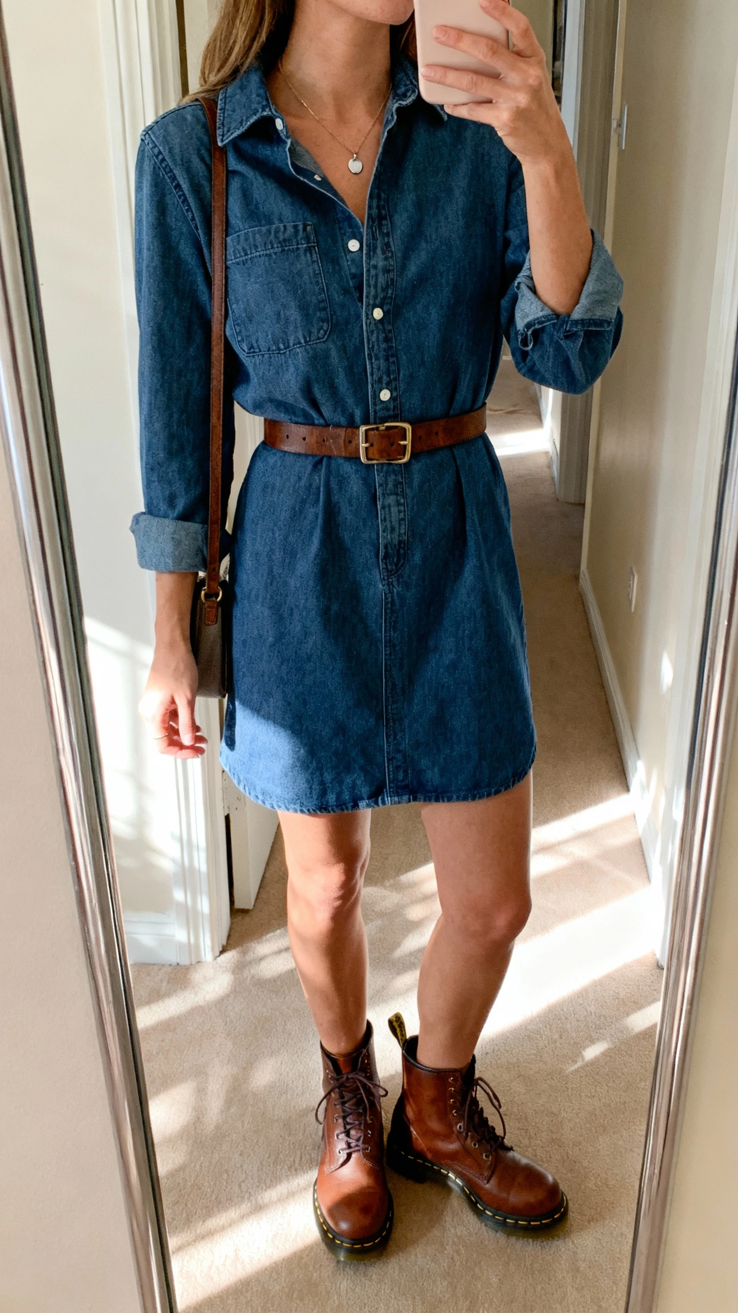 iPhone mirror selfie of a woman wearing brown Doc Martens with a denim shirt-dress, waist belt, subtle pendant necklace, and a small shoulder bag, face not visible, cropped at neck, entryway mirror with soft afternoon light, natural iPhone photo quality.