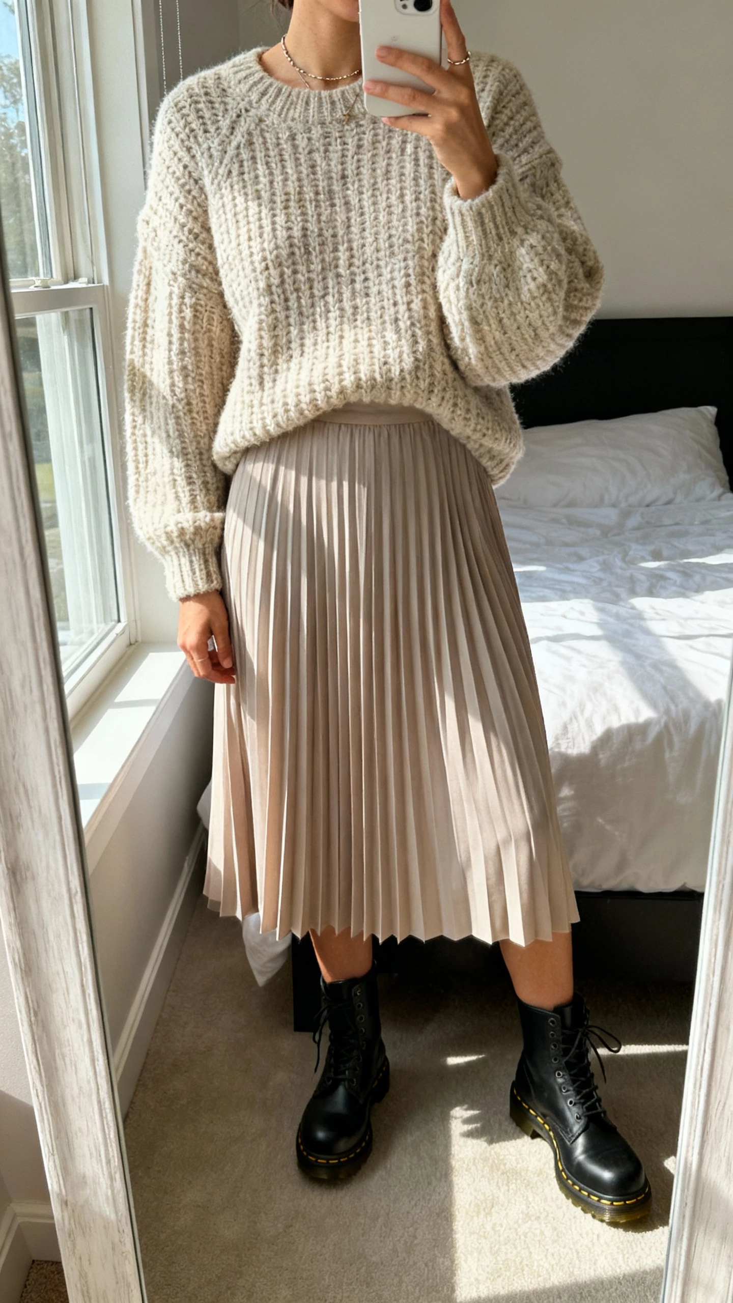 iPhone mirror selfie of a woman wearing chunky knit sweater, pleated midi skirt, Doc Martens combat boots, minimal jewelry, face not visible, cropped at neck, bedroom mirror with soft morning window light, natural iPhone photo quality.