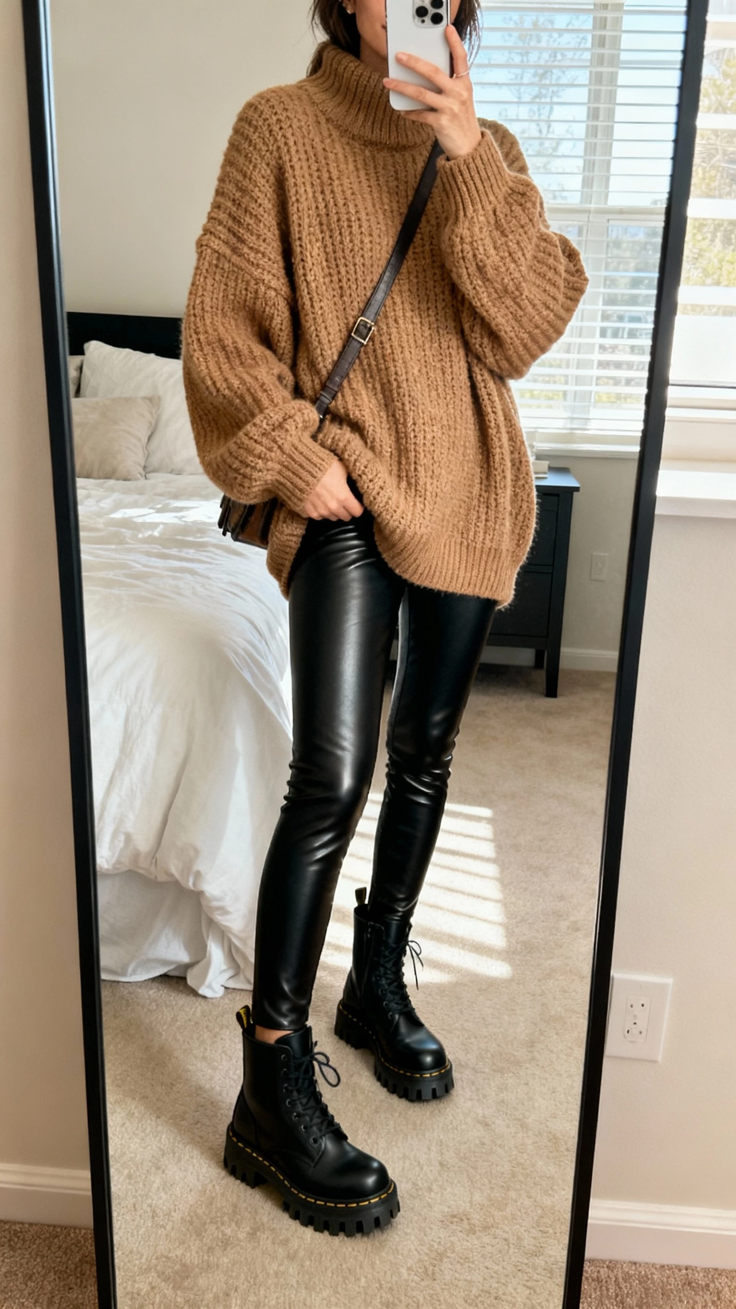 iPhone mirror selfie of a woman wearing faux-leather black leggings, oversized chunky knit sweater in tan, lug-sole ankle boots, crossbody bag, face not visible, bedroom full-length mirror, morning window light, natural iPhone quality
