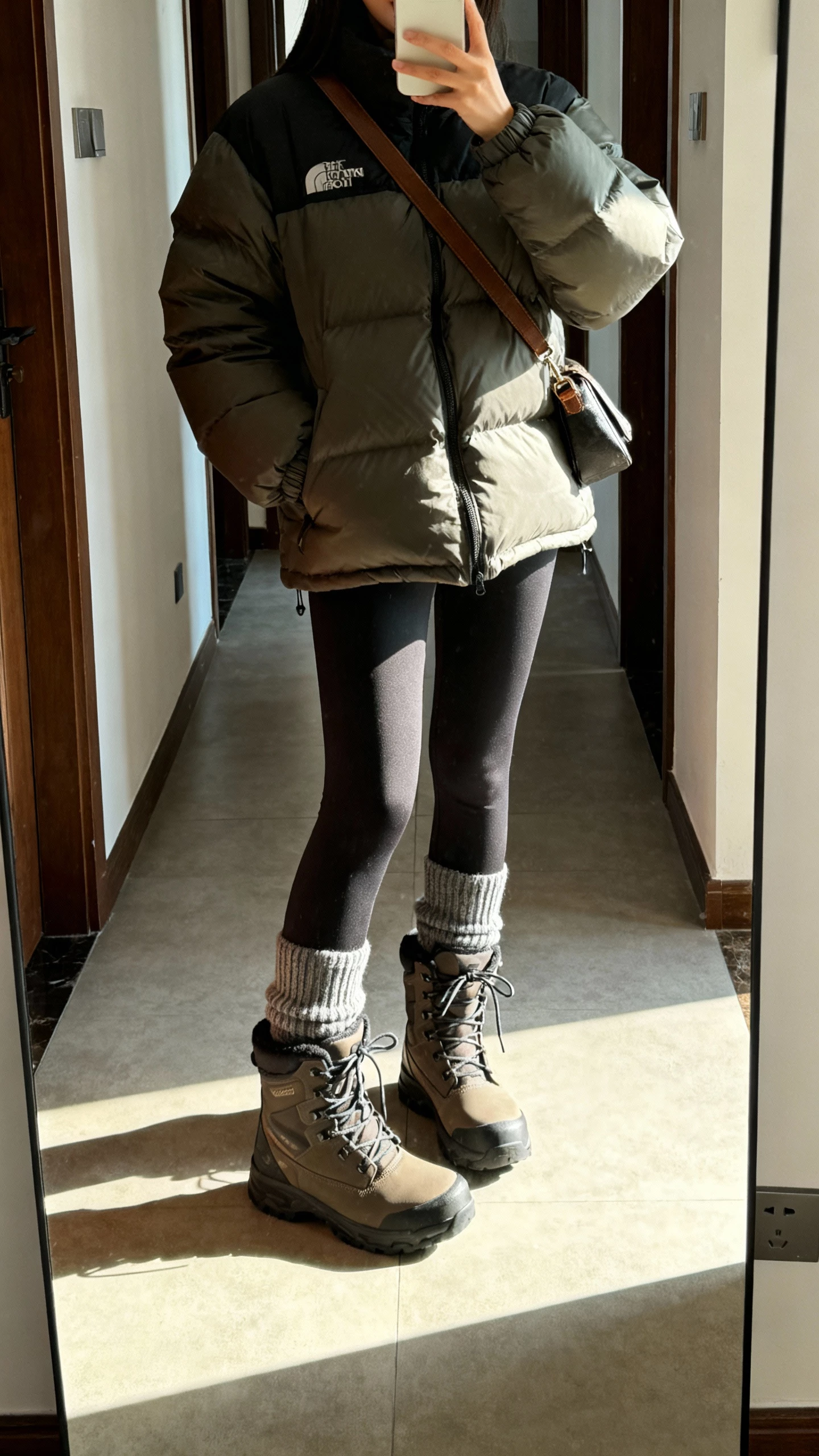 iPhone mirror selfie of a woman wearing insulated waterproof lace-up boots, puffer jacket, leggings, thick socks peeking above boots, crossbody bag, face not visible, apartment entryway mirror with daylight, natural iPhone aesthetic.