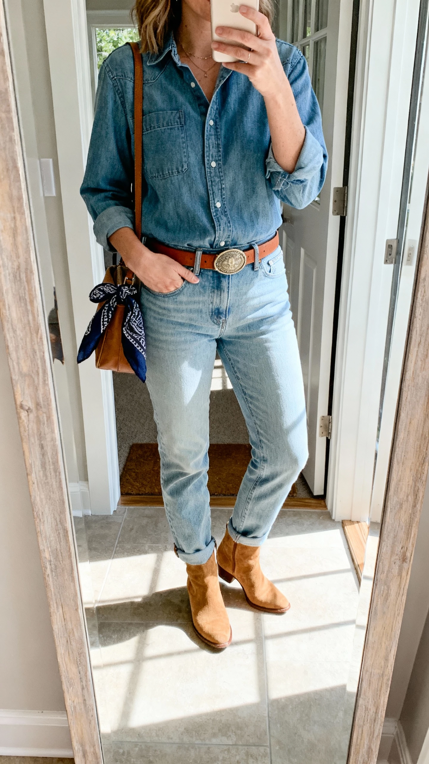 iPhone mirror selfie of a woman wearing light blue jeans with a denim shirt, tan suede ankle boots, slim brown belt, subtle concho-style buckle, bandana tied to bag, face not visible, entryway mirror with natural daylight, casual iPhone photo