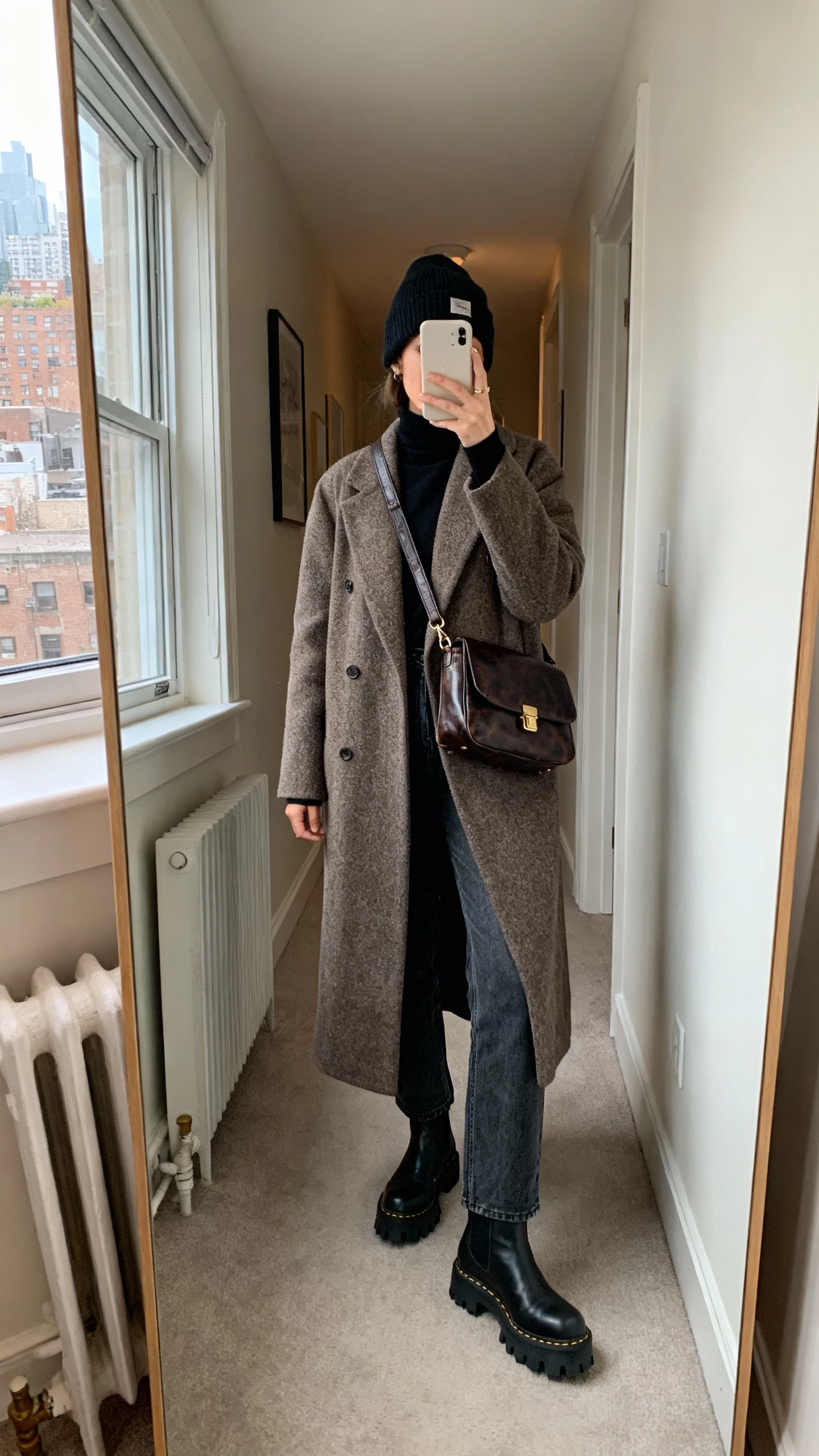 iPhone mirror selfie of a woman wearing lug-sole Chelsea boots with a long wool coat, black turtleneck, straight-leg jeans, leather crossbody, and beanie, face not visible, cropped at neck, city apartment hallway mirror, natural window light, natural iPhone photo quality.