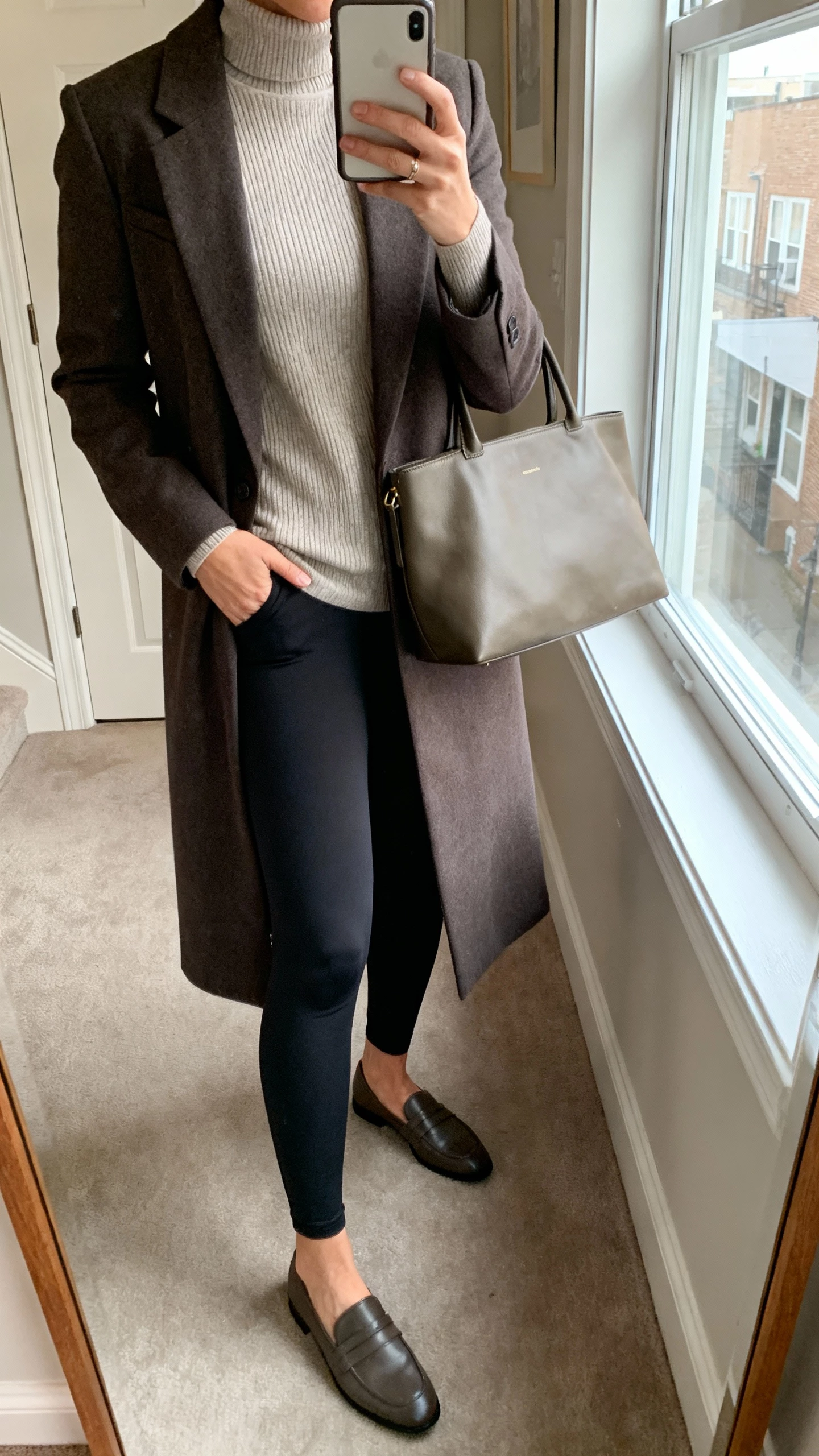 iPhone mirror selfie of a woman wearing ponte leggings with an elevated layered look: long blazer, fine-gauge turtleneck, loafers, structured tote, face not visible, apartment hallway mirror, morning window light, natural iPhone photo.