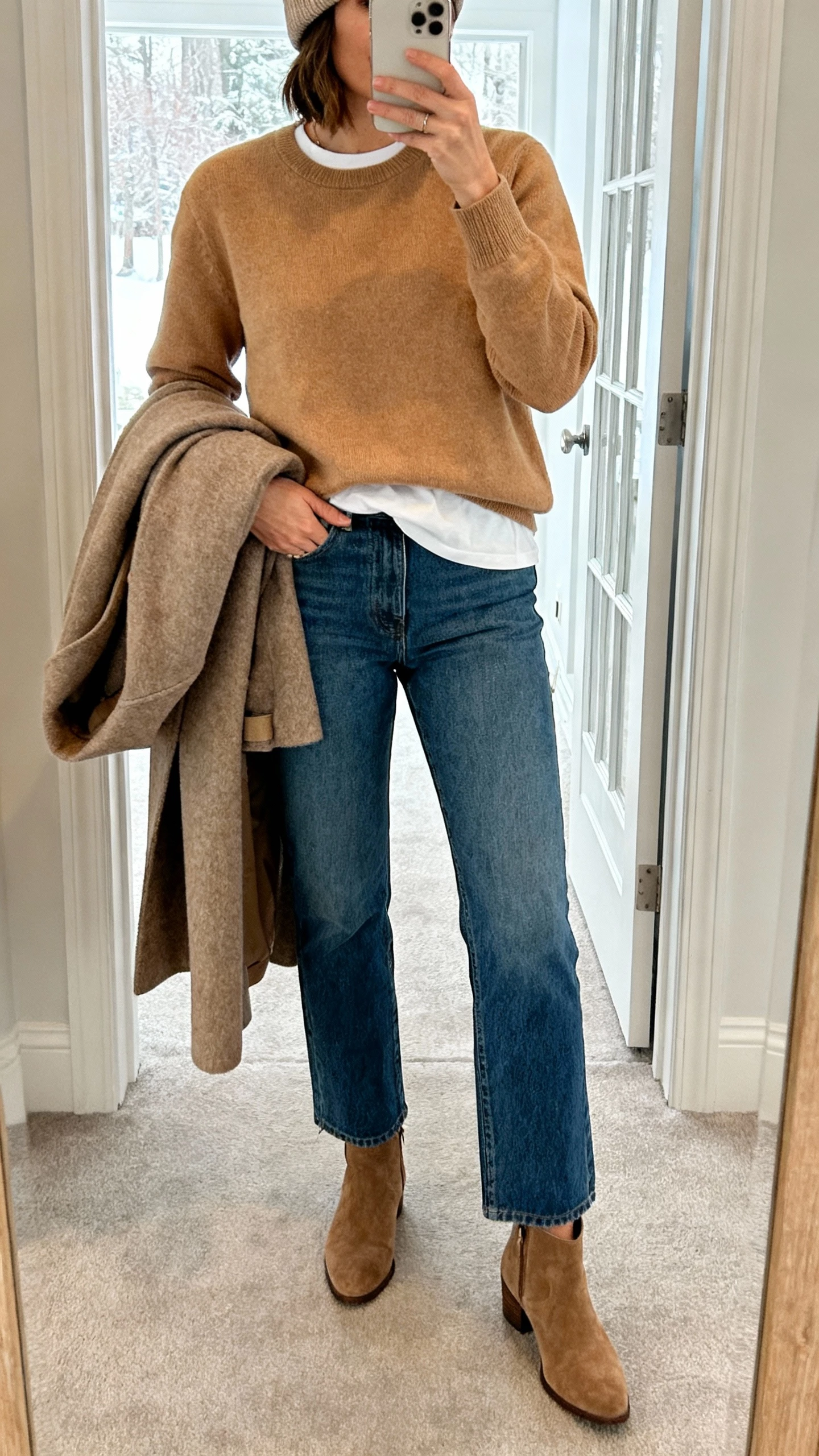 iPhone mirror selfie of a woman wearing straight-leg classic blue denim, camel cashmere crewneck layered over a white tee, wool coat draped over arm, suede ankle boots, beanie, face not visible, entryway mirror with soft winter light, natural iPhone photo.